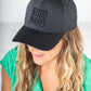 A person with long, wavy hair wears a comfortable black Emily McCarthy Signature Hat. Dressed in a green top, the individual gazes slightly downwards against a soft, neutral background.