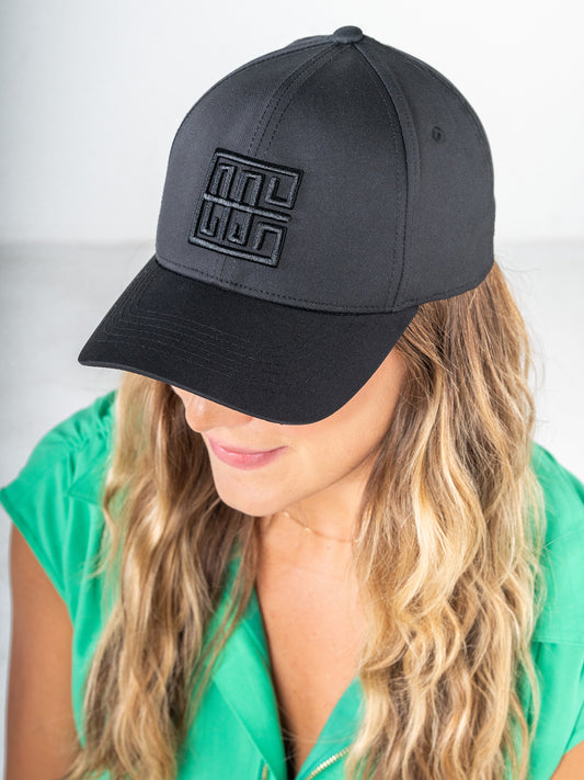 A person with long, wavy hair wears a comfortable black Emily McCarthy Signature Hat. Dressed in a green top, the individual gazes slightly downwards against a soft, neutral background.