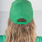 A person with long wavy blonde hair wears an Emily McCarthy Signature Hat in green, showcasing the signature embroidered on the back. Facing away, their shirt complements the hat against a plain, light-colored background.