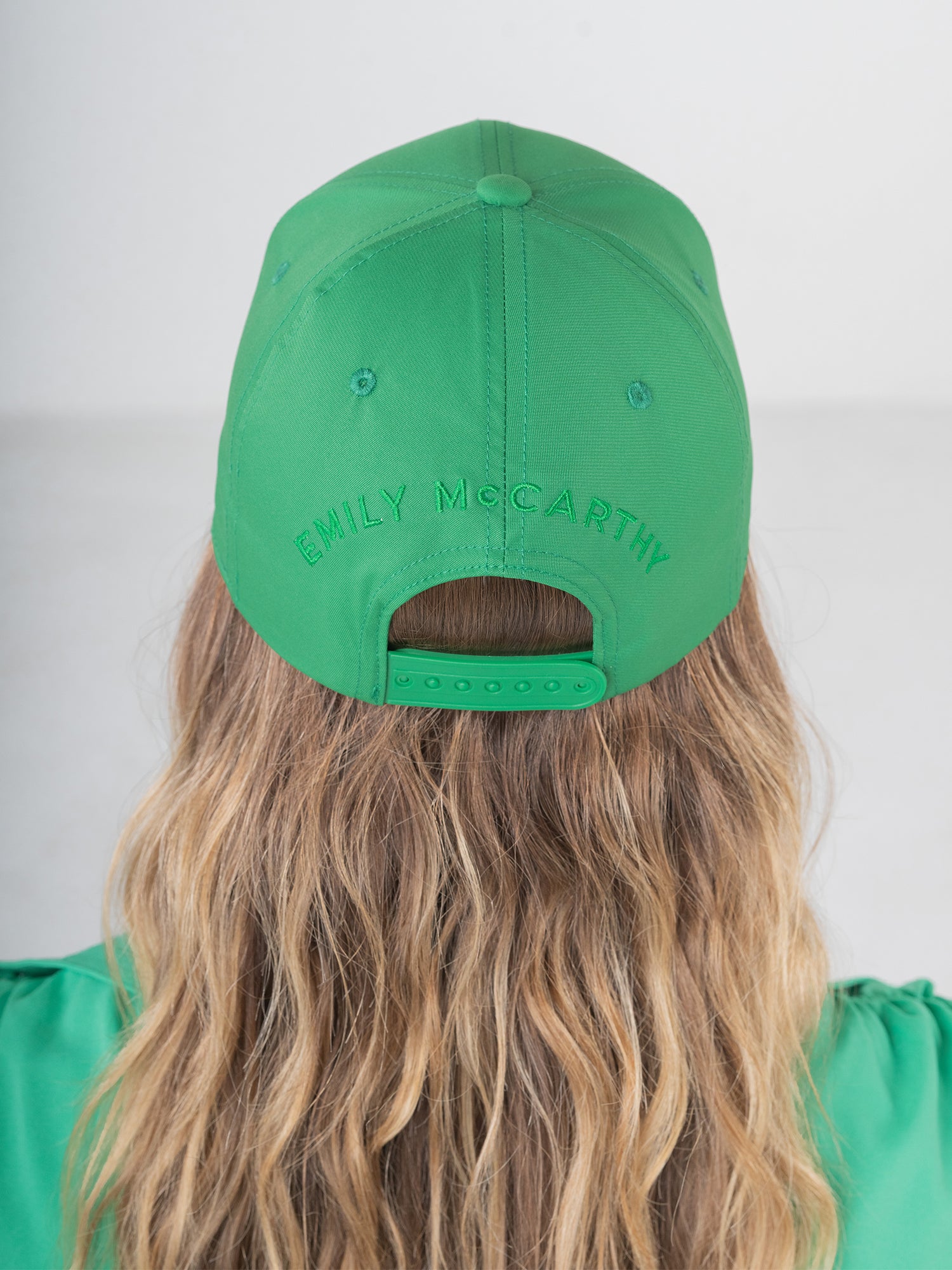 A person with long wavy blonde hair wears an Emily McCarthy Signature Hat in green, showcasing the signature embroidered on the back. Facing away, their shirt complements the hat against a plain, light-colored background.