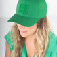 A woman with long, wavy blonde hair wears an Emily McCarthy Signature Hat in a vibrant green, perfectly complementing her sleeveless blouse. The hat, featuring a geometric logo for added style and comfort, enhances her fashionable look as she gazes downward against a neutral backdrop.