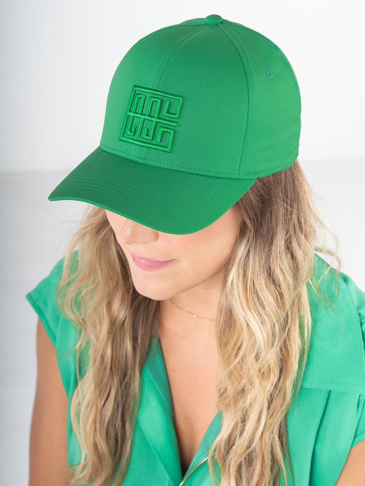 A woman with long, wavy blonde hair wears an Emily McCarthy Signature Hat in a vibrant green, perfectly complementing her sleeveless blouse. The hat, featuring a geometric logo for added style and comfort, enhances her fashionable look as she gazes downward against a neutral backdrop.