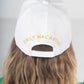 A person with long, wavy brown hair wears a white baseball cap from the Emily McCarthy Signature Hats collection, adorned with gold embroidery. The cap fits comfortably thanks to its adjustable strap and is paired with a green top against a light-colored background, making it perfect for post-workout attire.