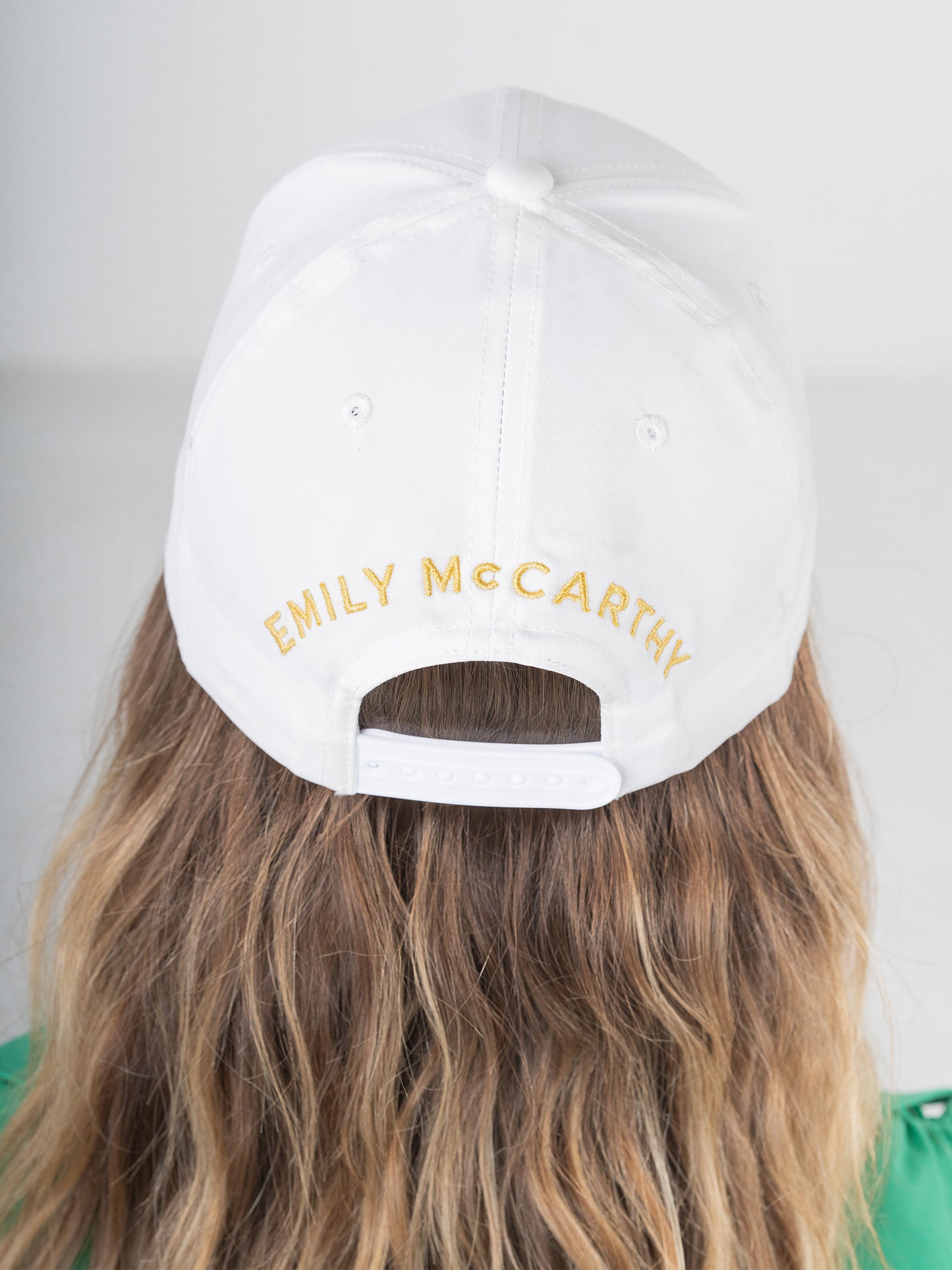 A person with long, wavy brown hair wears a white baseball cap from the Emily McCarthy Signature Hats collection, adorned with gold embroidery. The cap fits comfortably thanks to its adjustable strap and is paired with a green top against a light-colored background, making it perfect for post-workout attire.