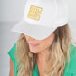 Emily McCarthy Signature hats provide both style and comfort, making them suitable for any event. In the scene, a person with long hair wears a white cap adorned with a yellow geometric design. Clad in a green top, they tilt their head slightly downwards against a simple light gray background, as if preparing for an exercise session.