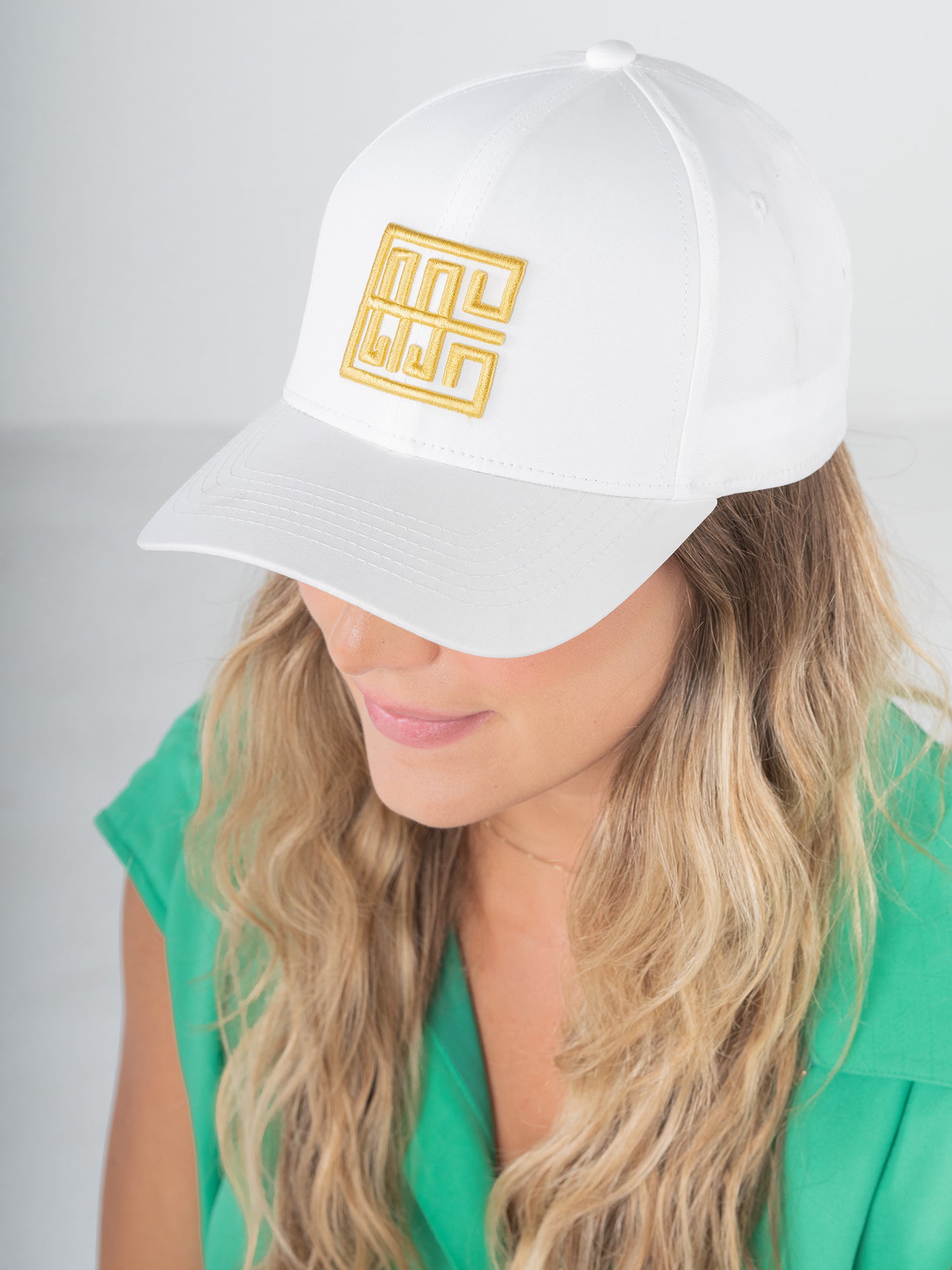 Emily McCarthy Signature hats provide both style and comfort, making them suitable for any event. In the scene, a person with long hair wears a white cap adorned with a yellow geometric design. Clad in a green top, they tilt their head slightly downwards against a simple light gray background, as if preparing for an exercise session.