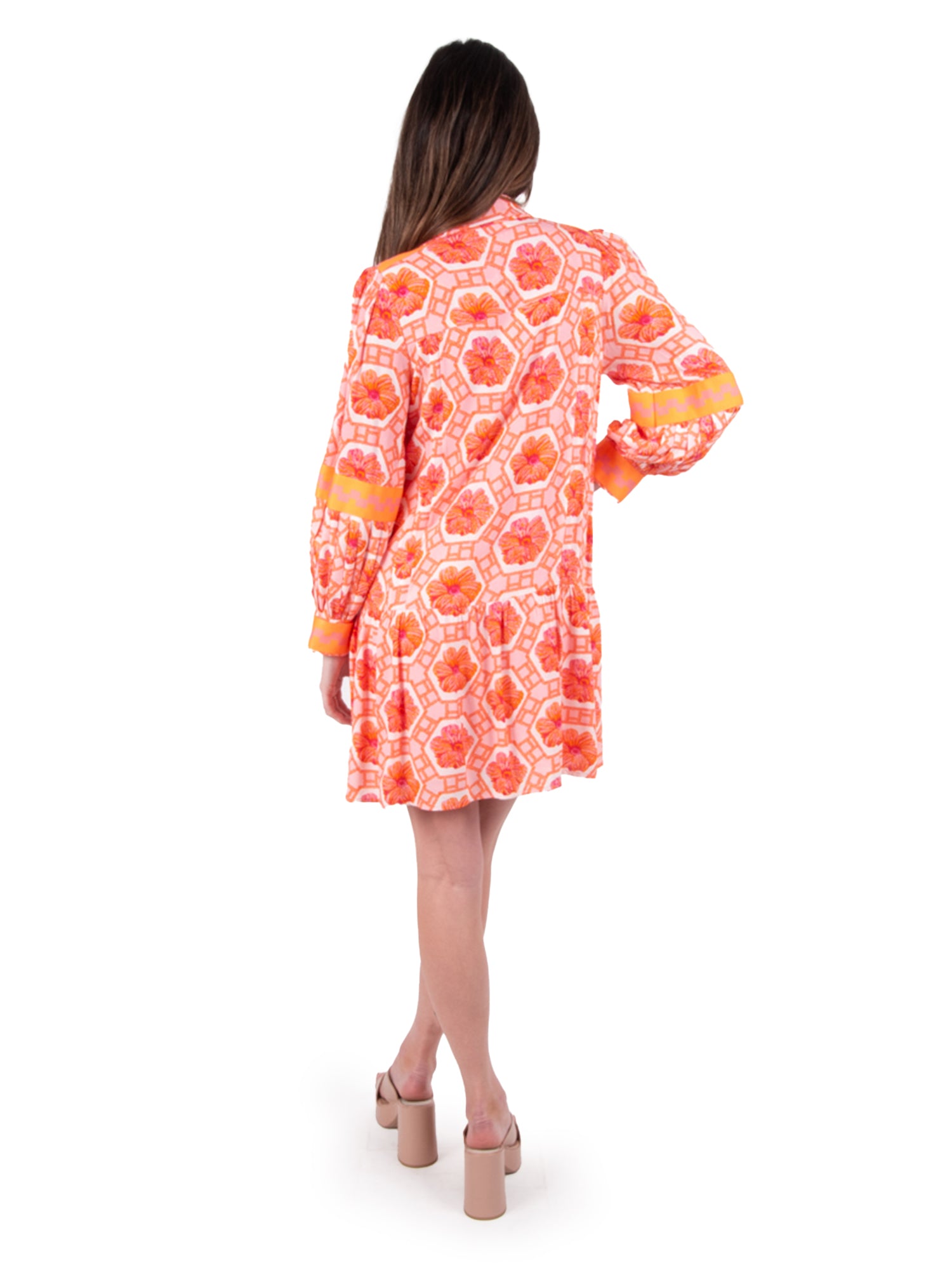 A person with long hair stands facing away, exuding modern elegance in a Floral Crochet Delany Dress featuring shades of orange and pink with long sleeves. High-heeled shoes complete the look against a white background, adding a vintage touch to their style.