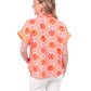 A person with long hair is wearing the Emily McCarthy Poppy Pullover - Floral Crochet, showcasing a vibrant floral hexagonal pattern in orange and pink shades. They pair it beautifully with white pants, inviting joy against a plain white backdrop.