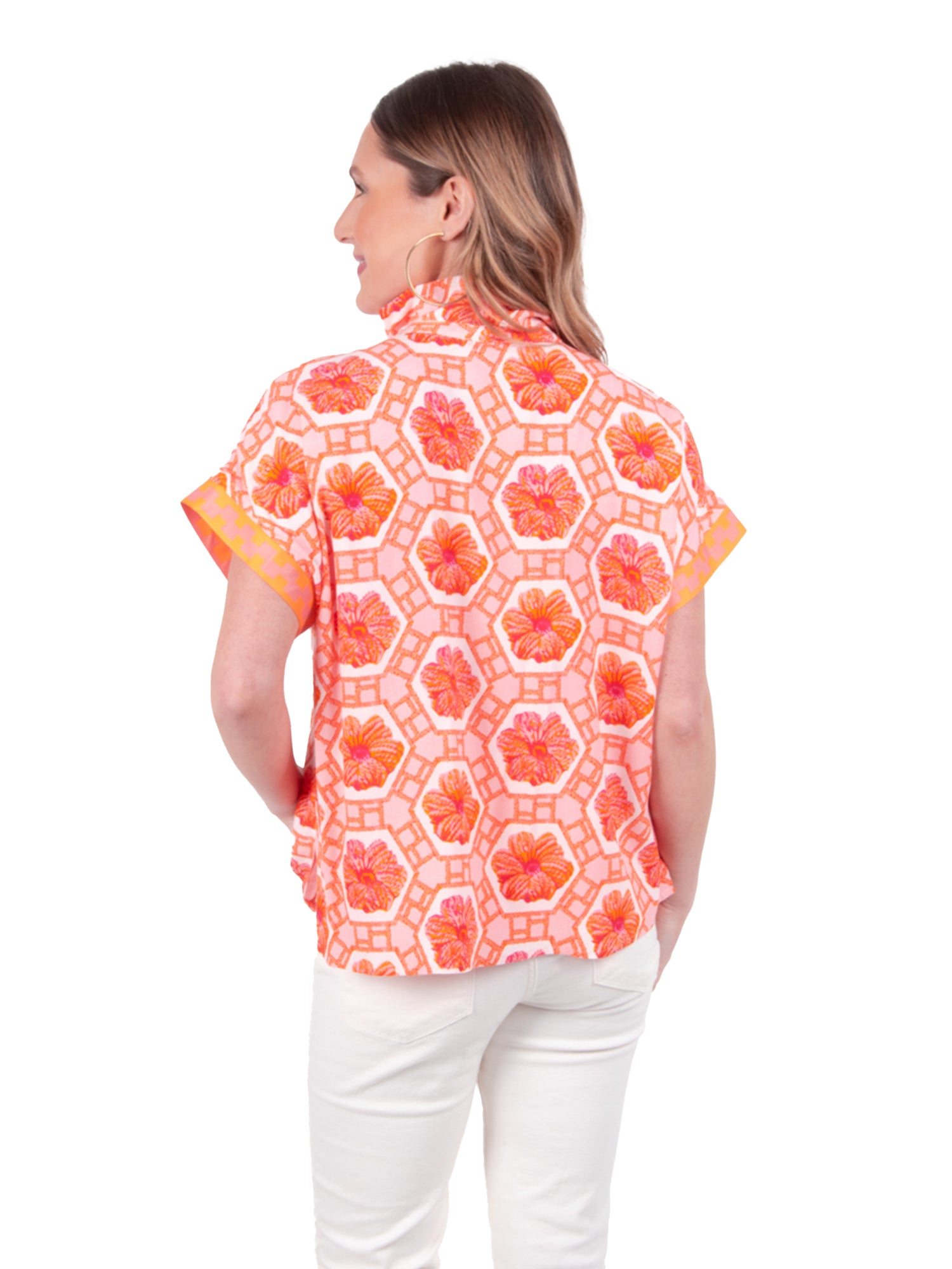 A person with long hair is wearing the Emily McCarthy Poppy Pullover - Floral Crochet, showcasing a vibrant floral hexagonal pattern in orange and pink shades. They pair it beautifully with white pants, inviting joy against a plain white backdrop.