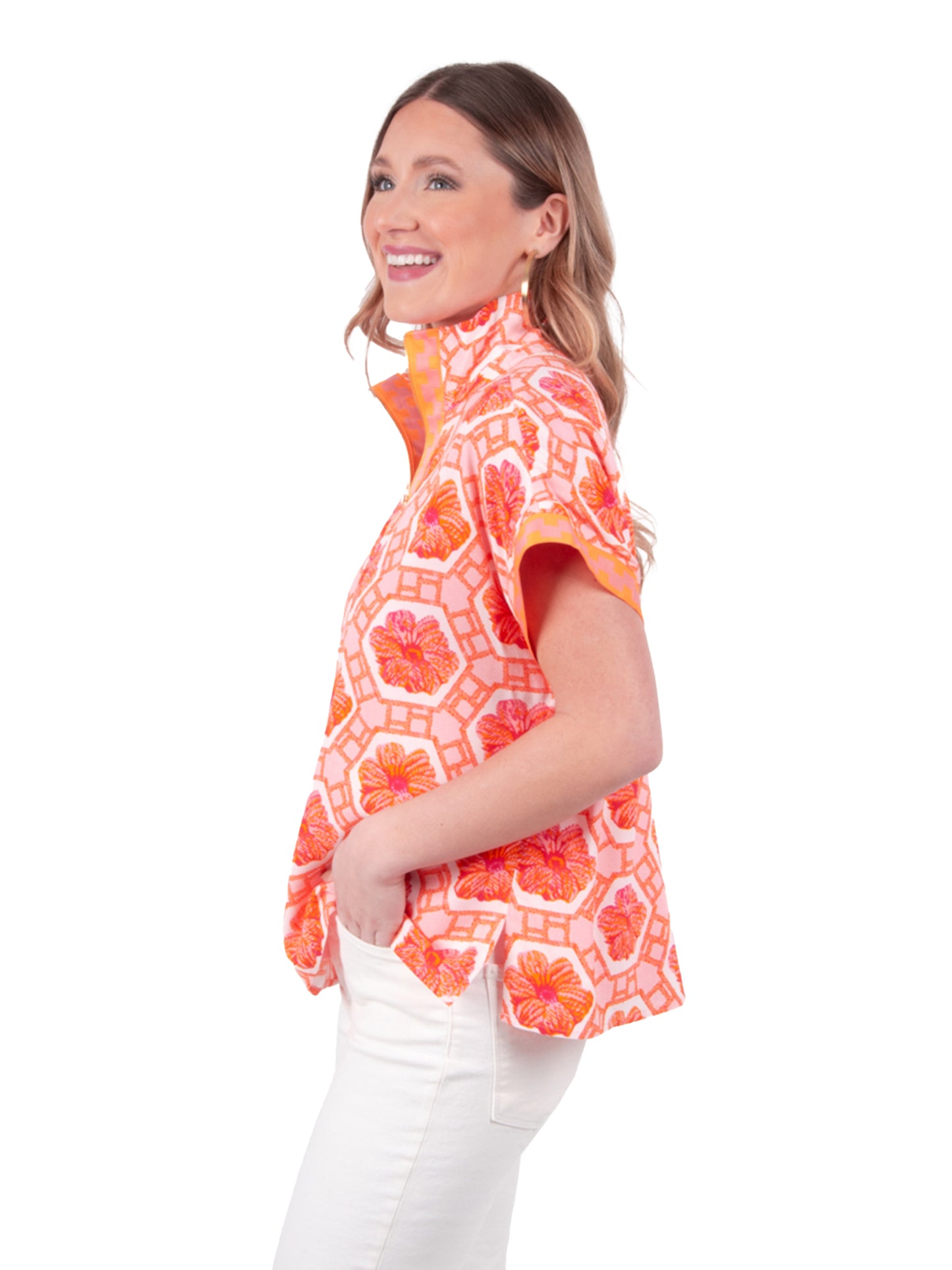 A woman wearing the Poppy Pullover – Floral Crochet blouse, featuring vibrant pink and orange patterns, a signature piece from Emily McCarthy Clothing, smiles while gazing to her left. With one hand tucked into the pocket of her white pants, she radiates an aura of joy against the plain white background.