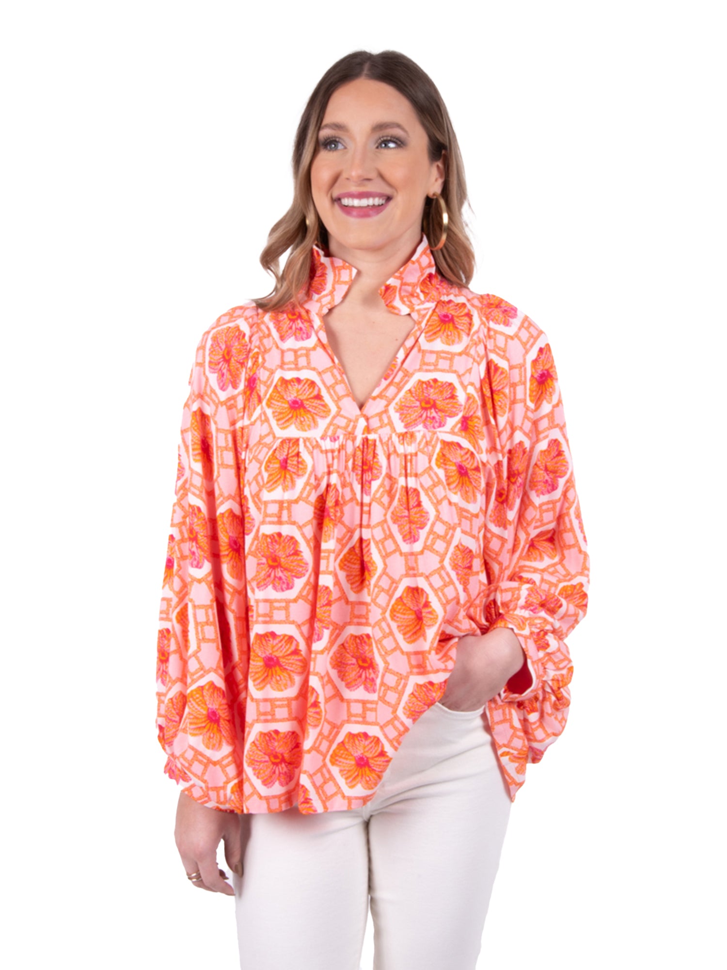 A person smiles while wearing the Stella Top - Floral Crochet, a brightly colored blouse adorned with orange and pink floral patterns. The top is designed with a ruffled collar and long sleeves. The individual has one hand in the pocket of their white pants, standing against a plain white backdrop.