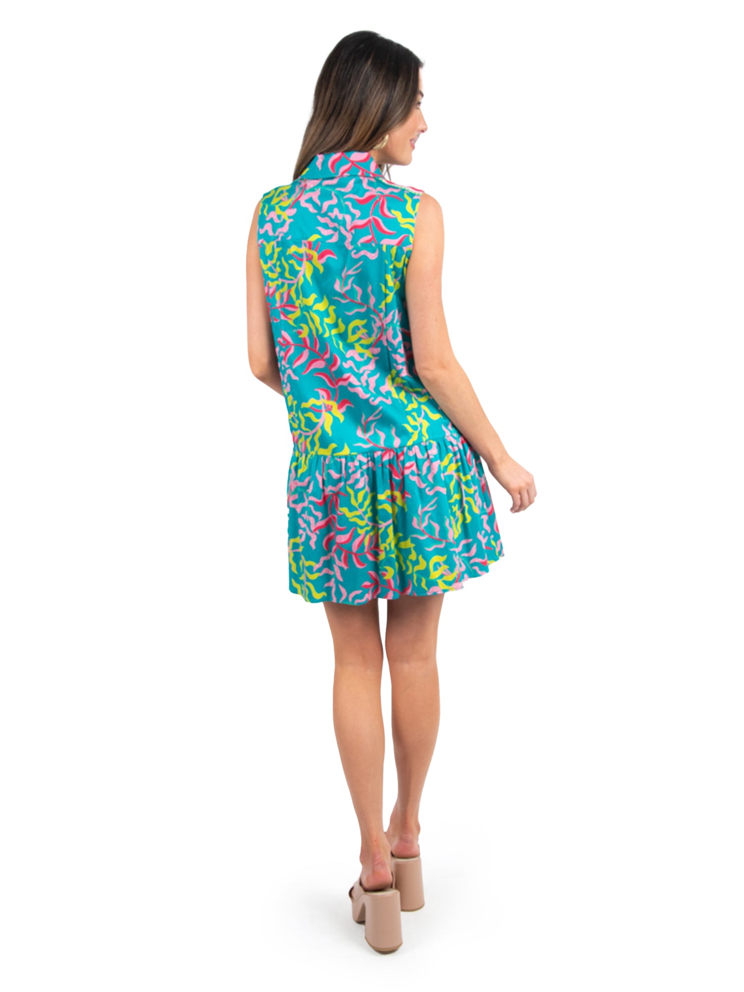 A woman in a sleeveless Daisy Dress - Floret stands facing away, highlighting its vibrant shades of blue, pink, and green. Her ensemble is both sporty and sophisticated with beige high-heeled sandals and long brown hair cascading down her back against a plain white background.