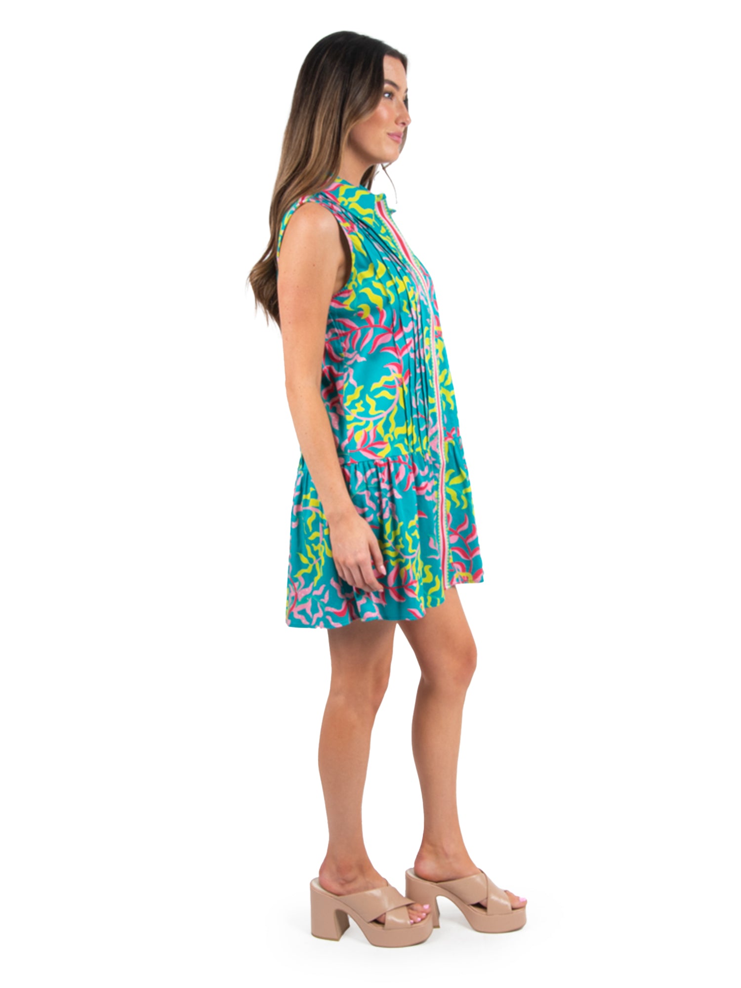 A woman stands in profile wearing the Daisy Dress - Floret, featuring abstract patterns in blue, pink, and yellow. She has long brown hair and is paired with nude platform sandals against a plain white backdrop, emphasizing her sporty and sophisticated style.