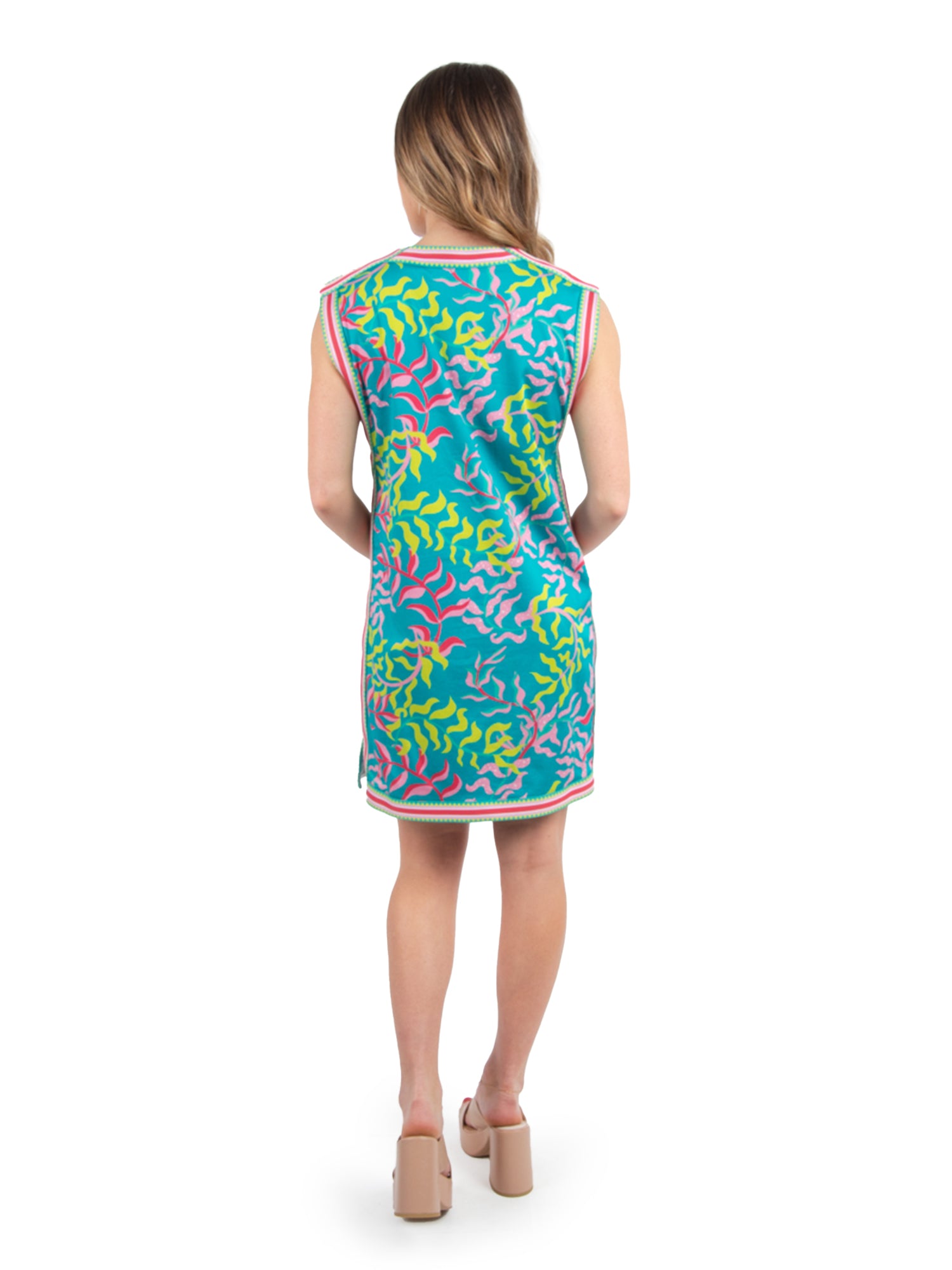 A woman wearing the Emily Shift Dress - Floret, a summer staple featuring colorful, abstract patterns in vibrant pink, teal, and yellow. The sleeveless, knee-length dress offers a versatile look when paired with beige block-heeled sandals against a plain white background.