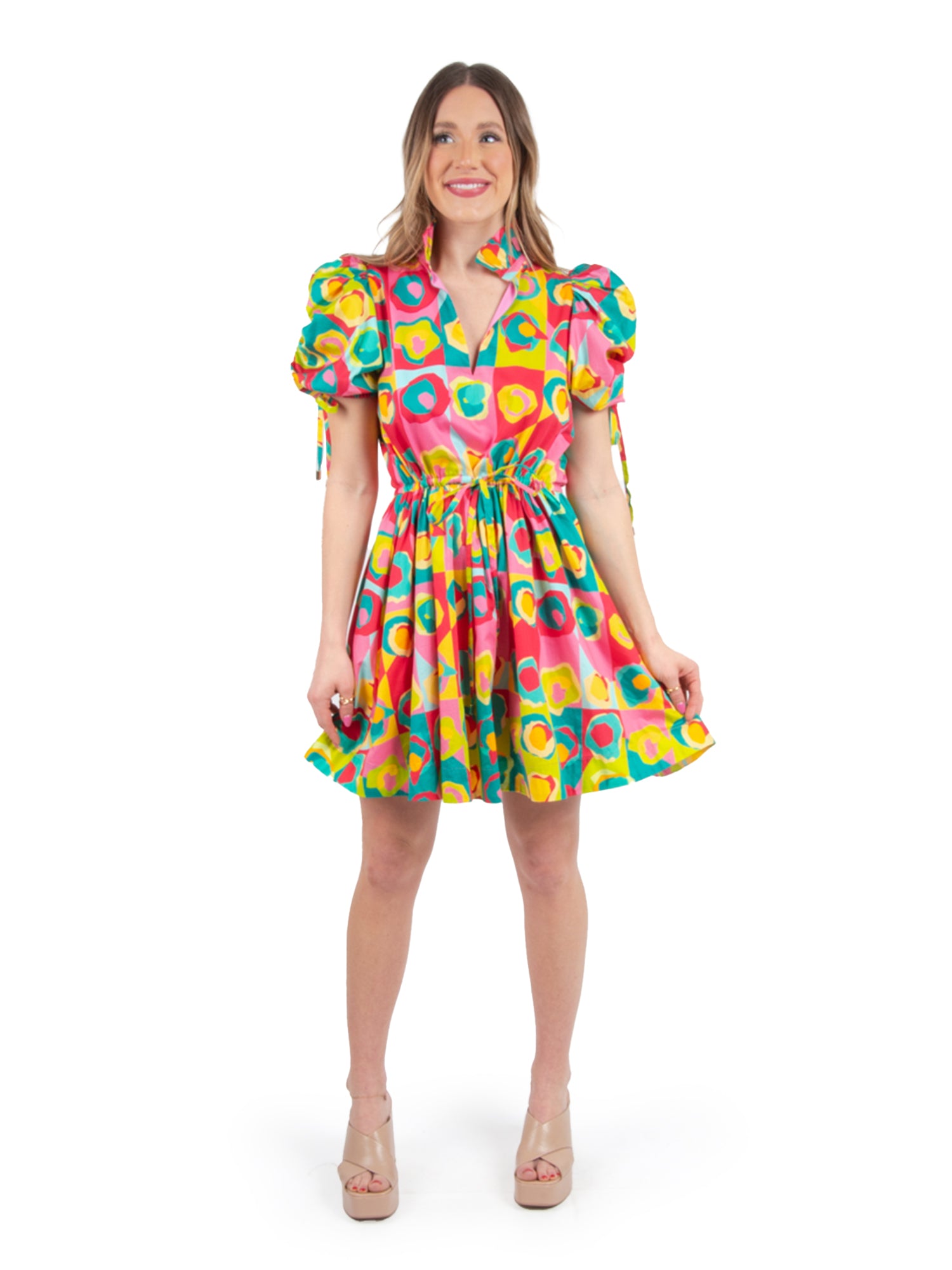 A woman wearing the colorful Posey Dress - Rhett Pop, adorned with geometric patterns, poses with a smile. The dress, highlighted by its puff sleeves and flared skirt, exudes a chic design. She pairs it with beige high heels while standing against a plain white background.