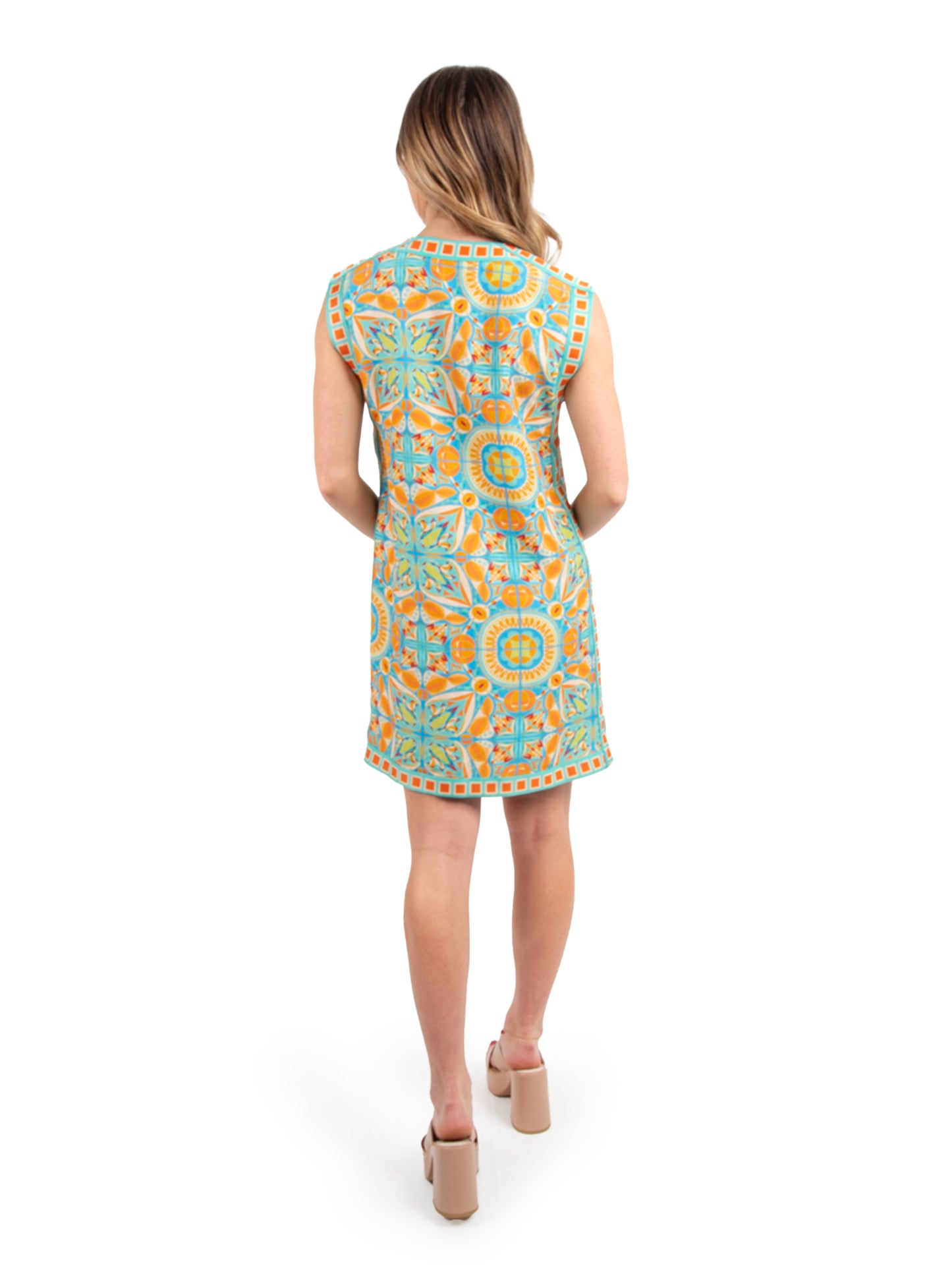 A woman in an Emily Shift Dress - Poolside stands with her back to the camera, showcasing the sleeveless dress from the Emily McCarthy Clothing line with its vibrant Amalfi Coast print featuring geometric designs in blue, orange, and yellow. She accessorizes her outfit with elegant beige heels.