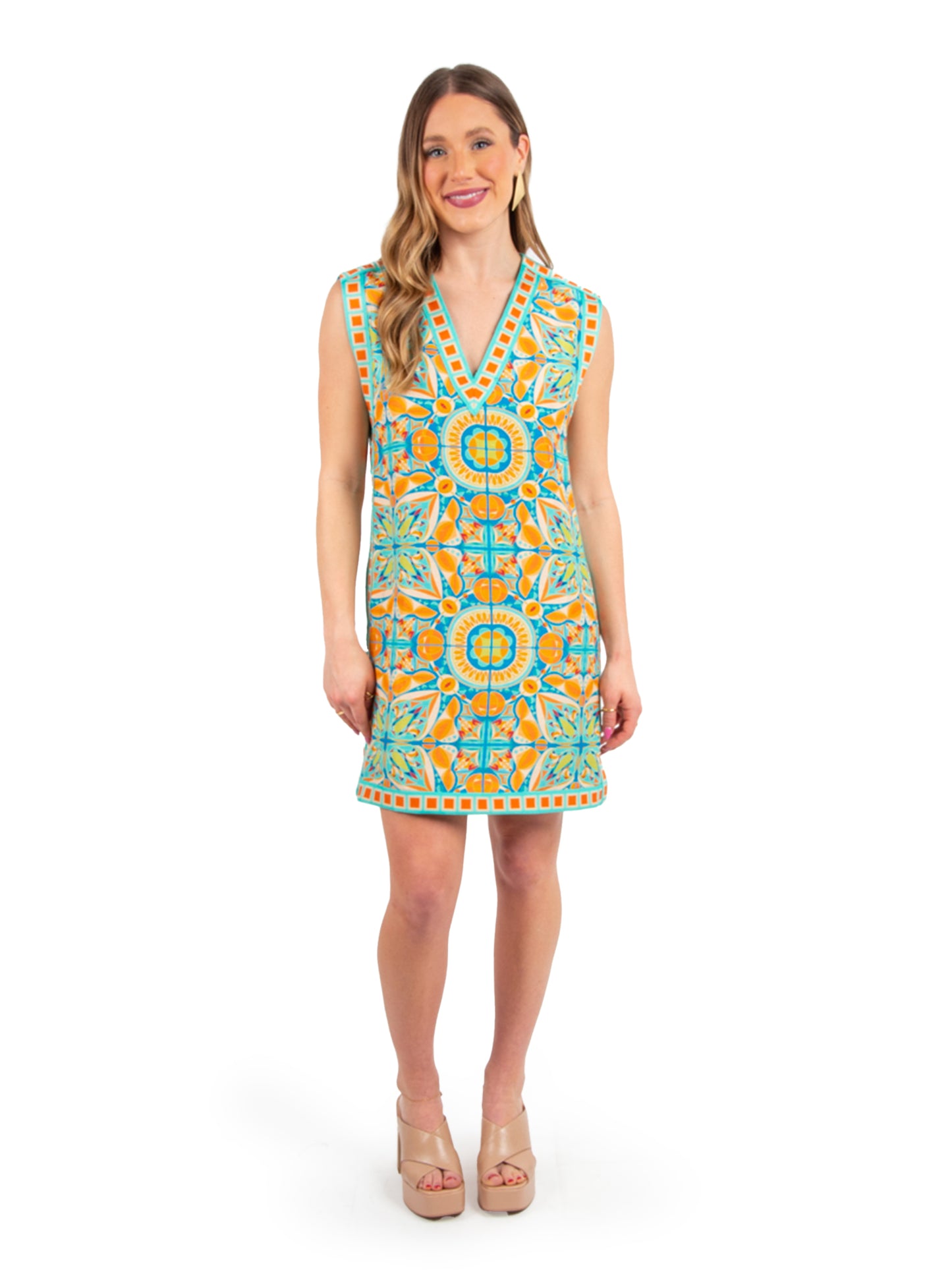 A woman in an Emily McCarthy Clothing Emily Shift Dress - Poolside stands smiling against a white background. Her colorful, patterned sleeveless dress features vibrant geometric designs in orange and blue. She has long, wavy hair and is wearing nude platform sandals.