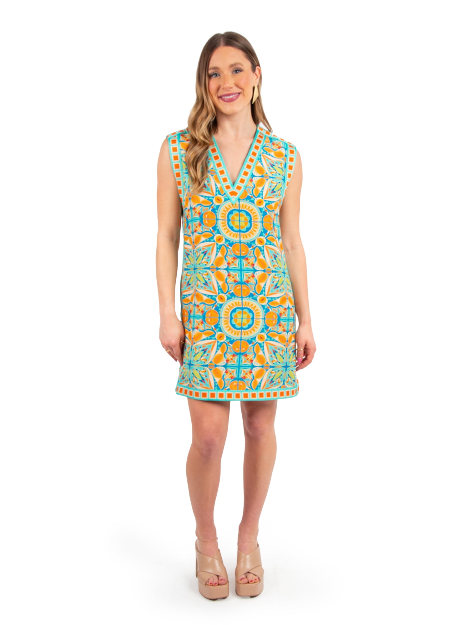 A woman in an Emily McCarthy Clothing Emily Shift Dress - Poolside stands smiling against a white background. Her colorful, patterned sleeveless dress features vibrant geometric designs in orange and blue. She has long, wavy hair and is wearing nude platform sandals.