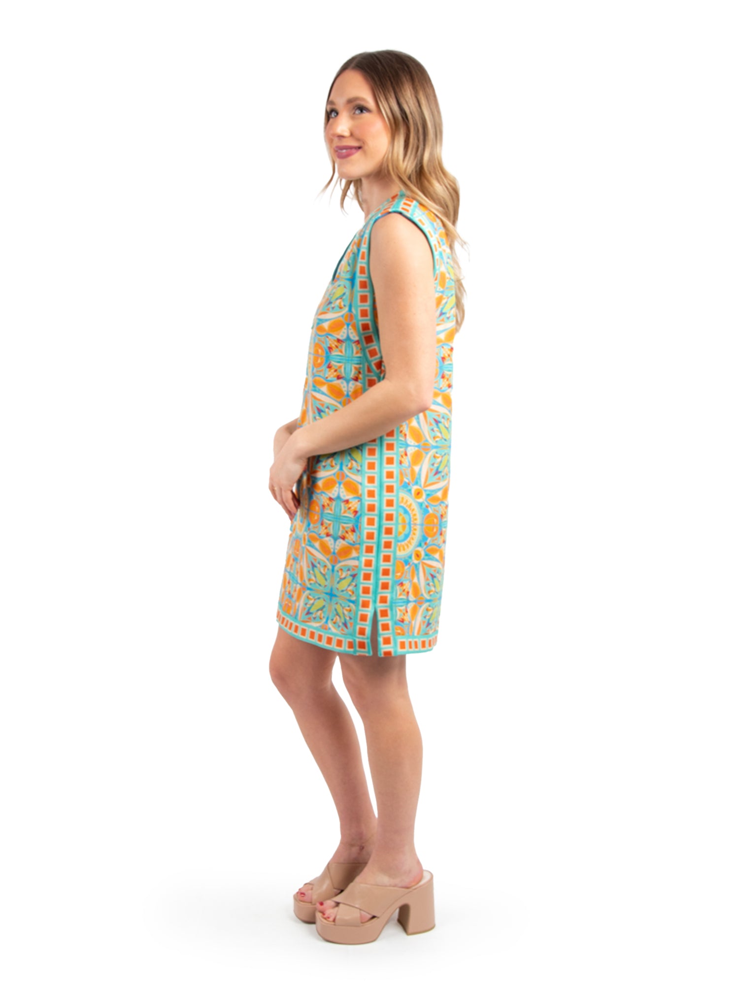 A woman stands sideways in an Emily Shift Dress - Poolside, showcasing a vibrant Amalfi Coast print with hues of blue, orange, and green. She pairs it with nude platform heels and has long, wavy hair. Her gentle smile is set against a plain white background.