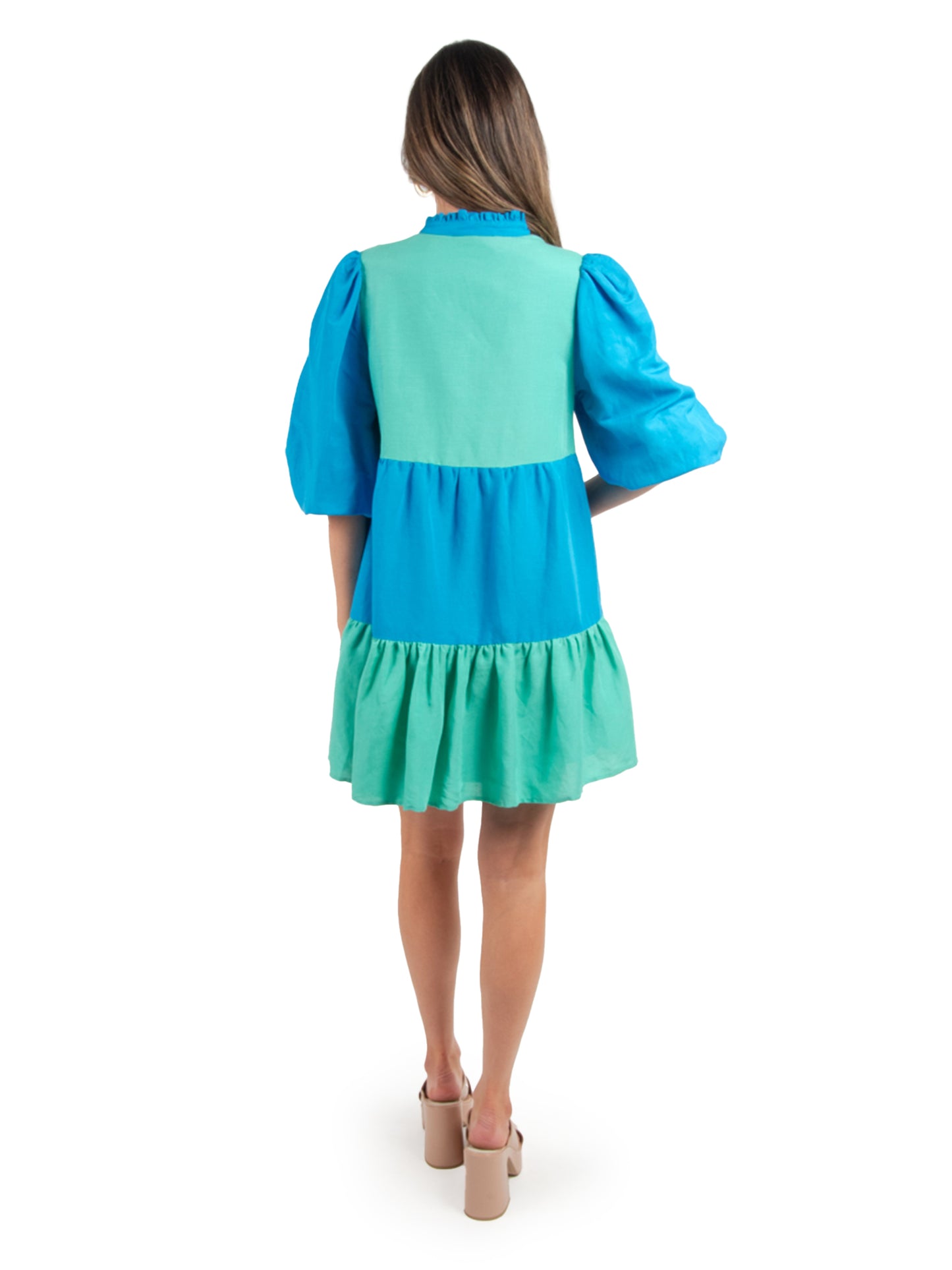 A person with long hair is shown from behind in a Frankie Dress - Lakeside Colorblock, which features puffed sleeves in shades of blue and green. They stand against a plain white background, wearing beige block-heeled sandals.