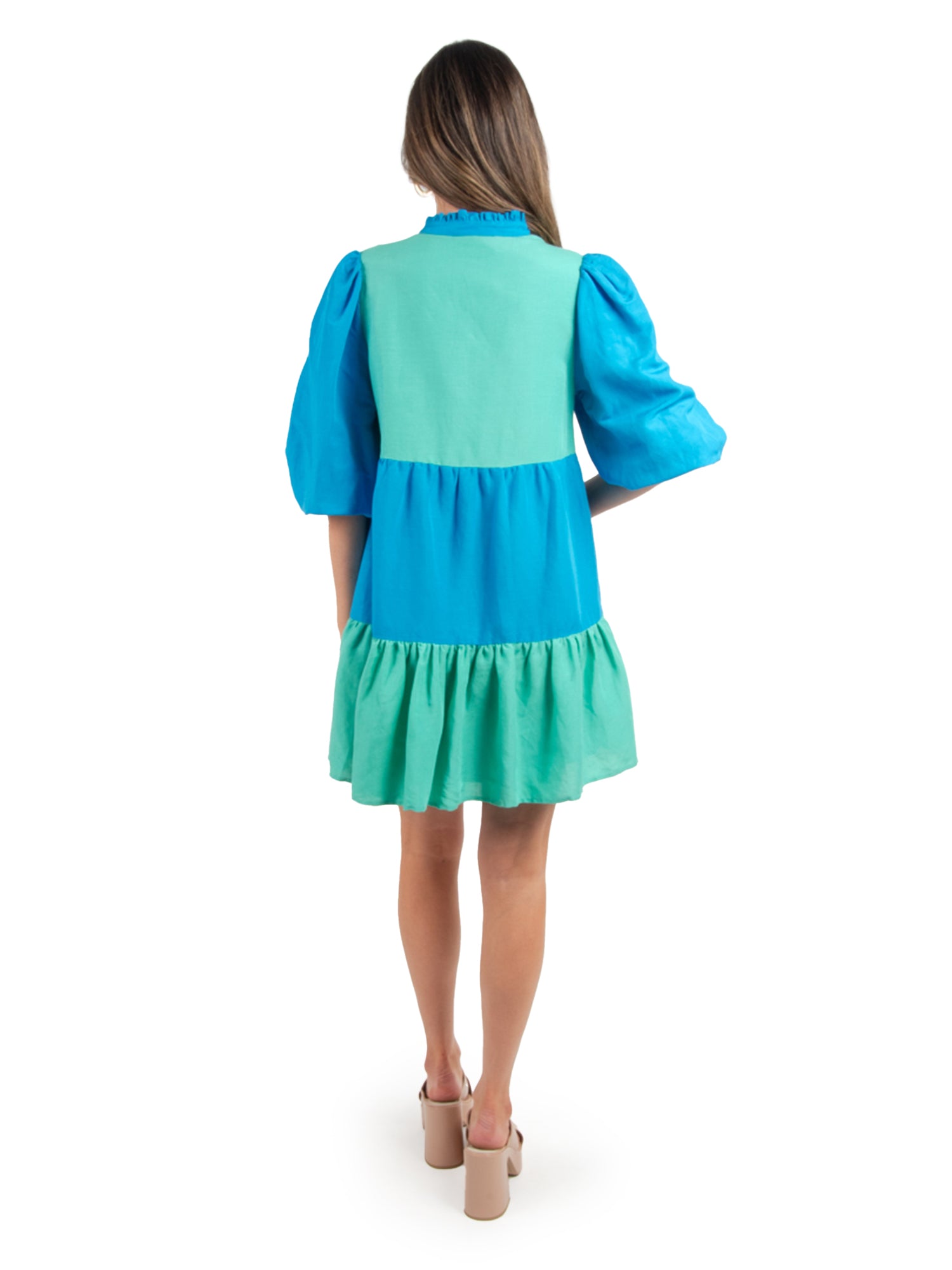 A person with long hair is shown from behind in a Frankie Dress - Lakeside Colorblock, which features puffed sleeves in shades of blue and green. They stand against a plain white background, wearing beige block-heeled sandals.