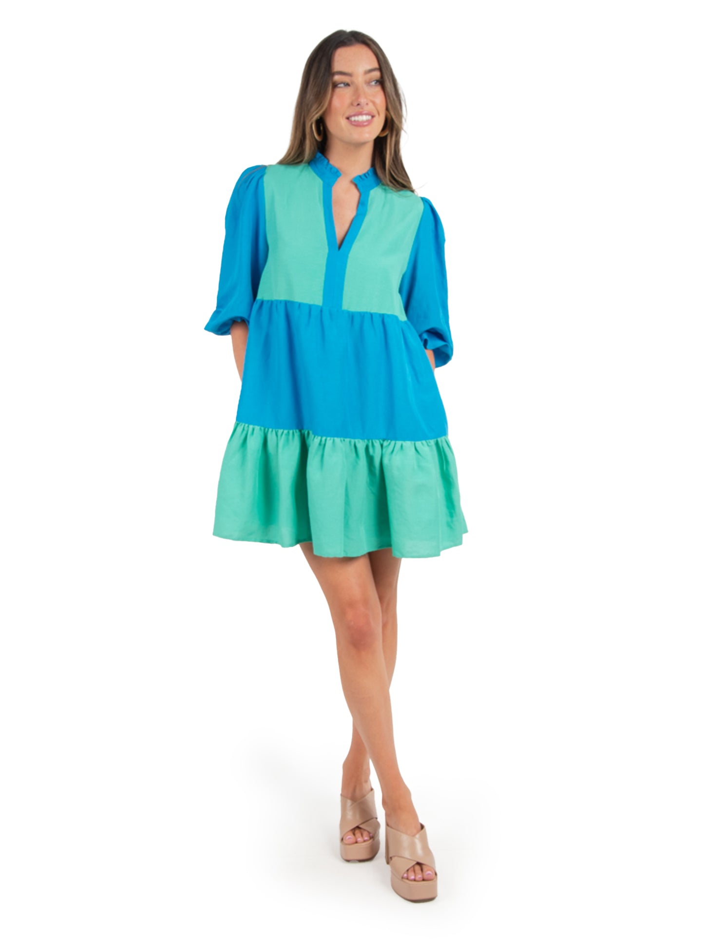 A person wearing a vibrant "Frankie Dress - Lakeside Colorblock" with a v-neck and puffed sleeves stands smiling. The knee-length dress showcases bright blue and teal hues. They have long hair and are wearing beige platform shoes against a plain white background.