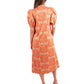 A woman stands with her back to the camera, showcasing the orange-patterned Hampton Dress - Cloister by Emily McCarthy, which features puff sleeves. Her long hair flows freely down her shoulders. She pairs the ensemble with beige platform sandals against a simple white backdrop.