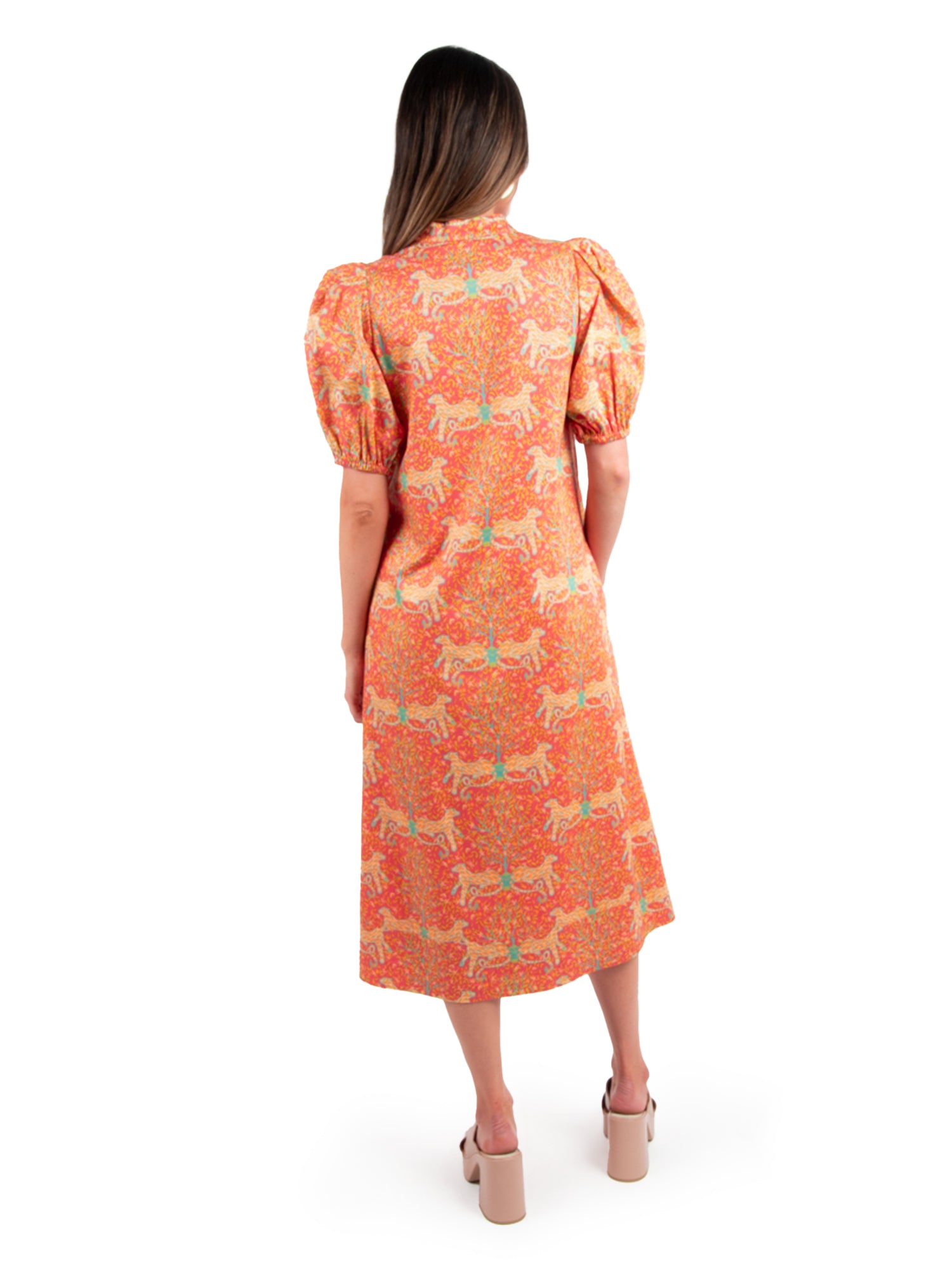 A woman stands with her back to the camera, showcasing the orange-patterned Hampton Dress - Cloister by Emily McCarthy, which features puff sleeves. Her long hair flows freely down her shoulders. She pairs the ensemble with beige platform sandals against a simple white backdrop.