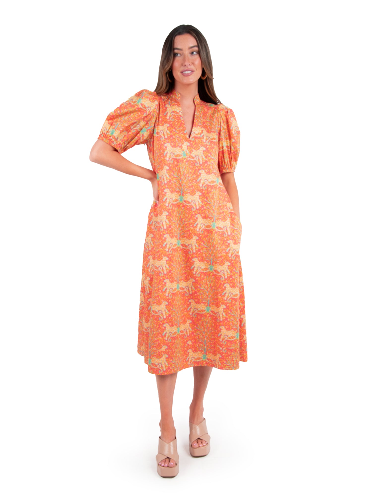 A person stands wearing the Hampton Dress - Cloister by Emily McCarthy, which features a horse pattern along with a V-neckline and puff sleeves. They have long brown hair and are styled with pale platform sandals against a white background.