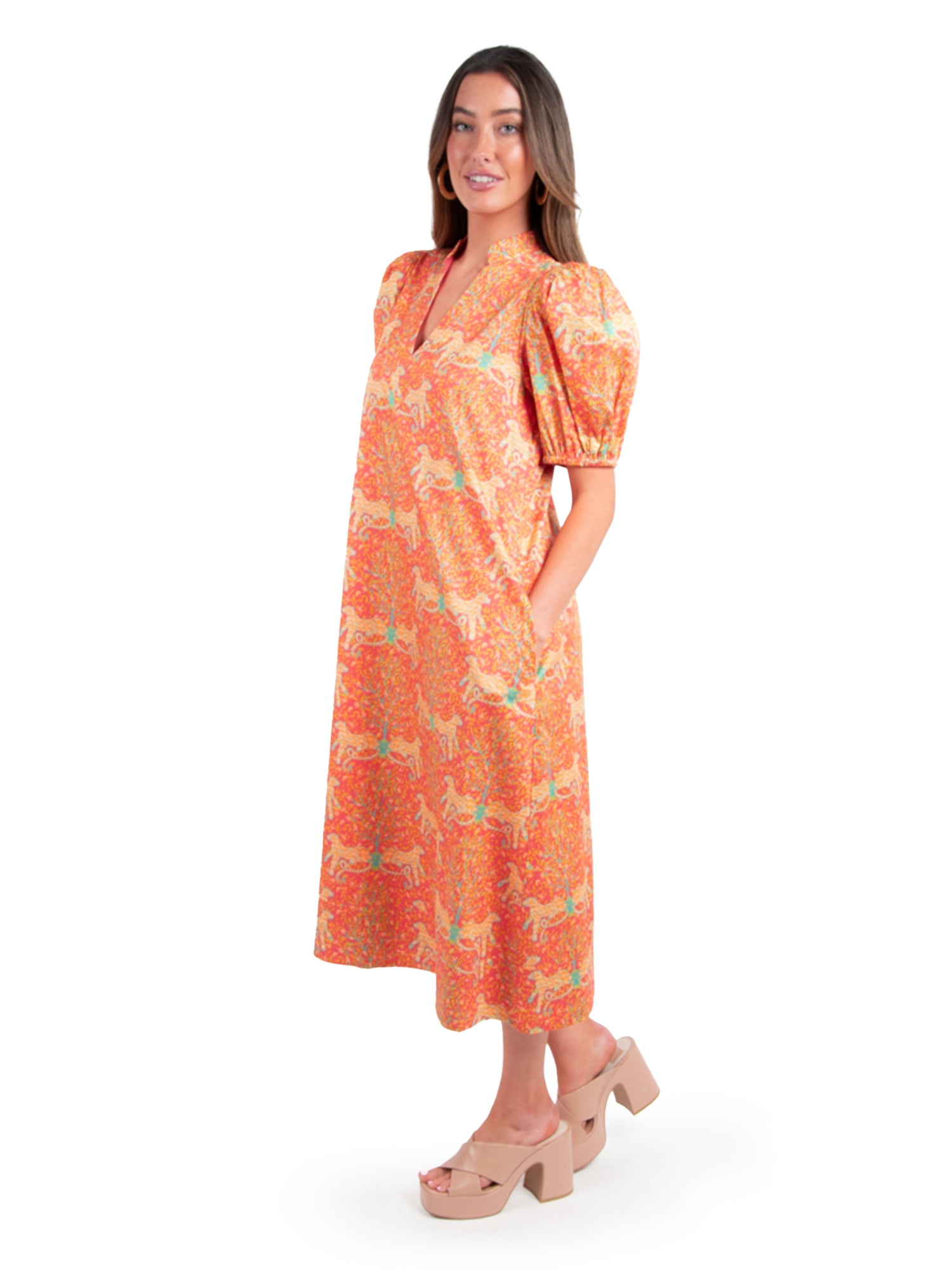 A woman stands wearing the Hampton Dress - Cloister by Emily McCarthy, in vibrant orange and adorned with a colorful pattern of horses. The dress features short puff sleeves and a V-neckline. She complements her look with platform sandals and poses confidently with one hand in her dress pocket against a plain white background.