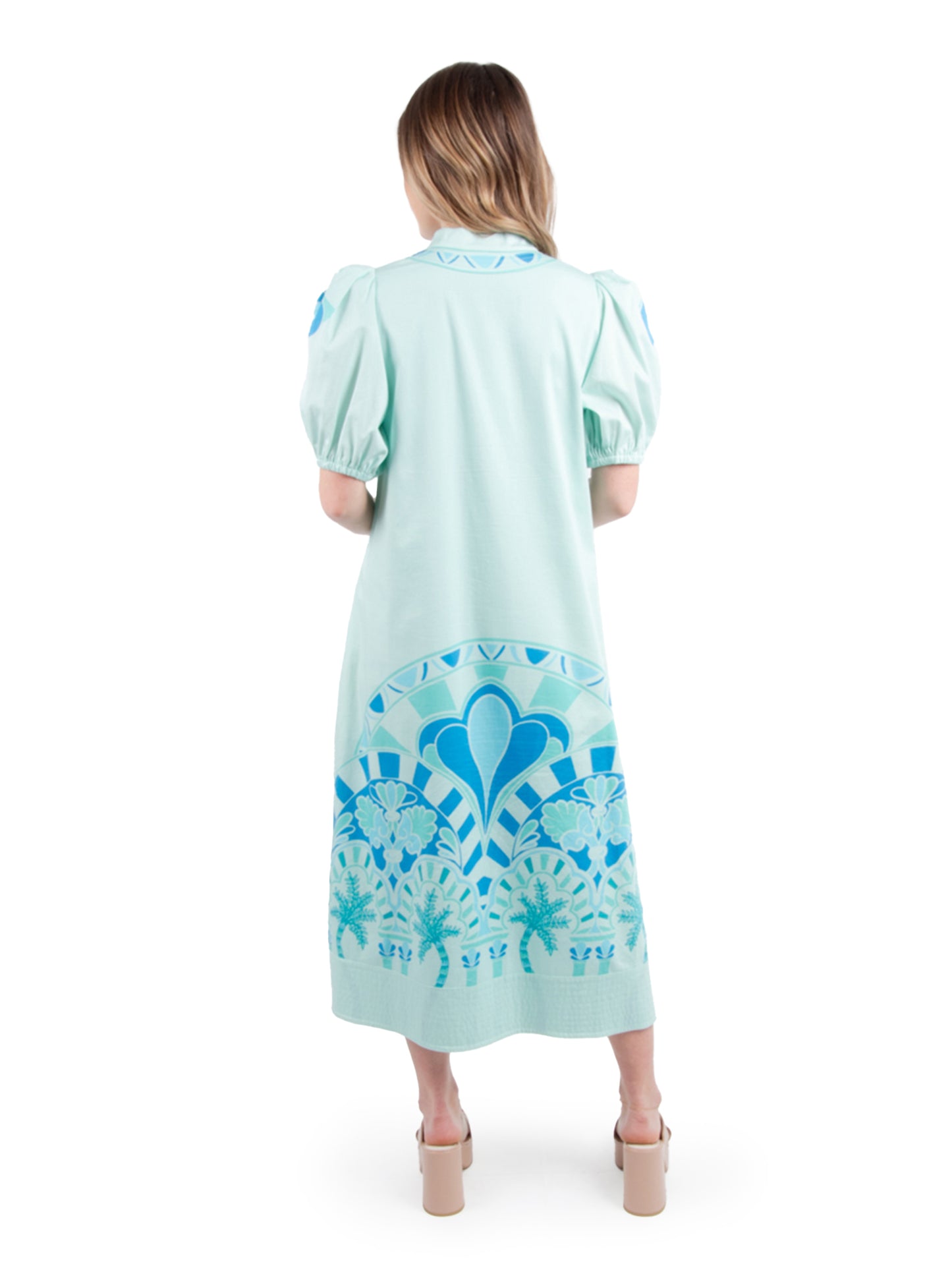 Back view of a woman in the Hampton Dress - Cordoba, showcasing its blue geometric and floral patterns. This dress features short puff sleeves and is styled with beige heeled sandals against a white background, highlighting its versatility for any occasion.