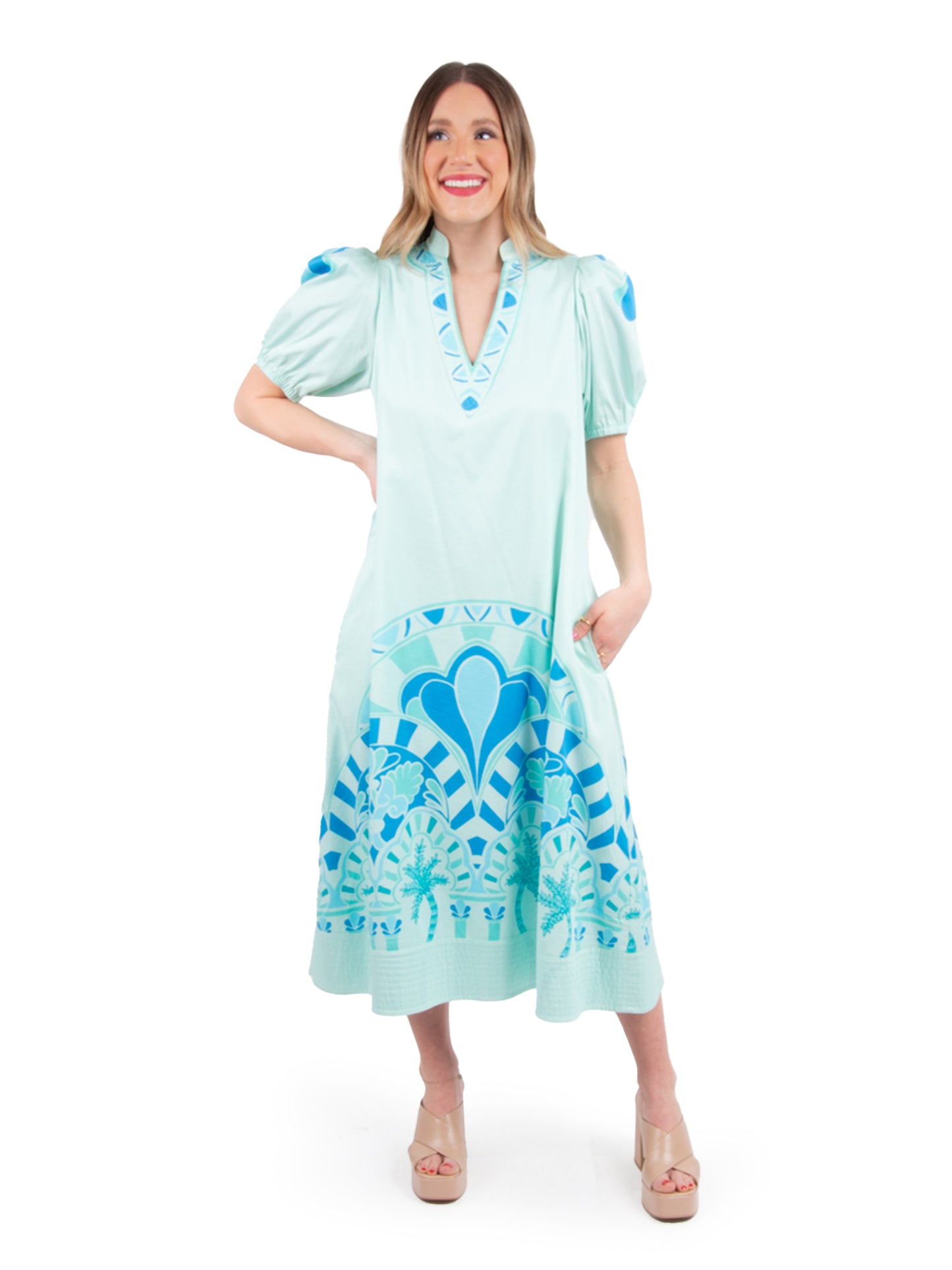 A woman smiles in the light teal Hampton Dress - Cordoba, featuring puff sleeves and blue geometric patterns, along with convenient pockets. She stands confidently with a hand on her hip against a white background, showcasing the versatile styling of her beige platform sandals.