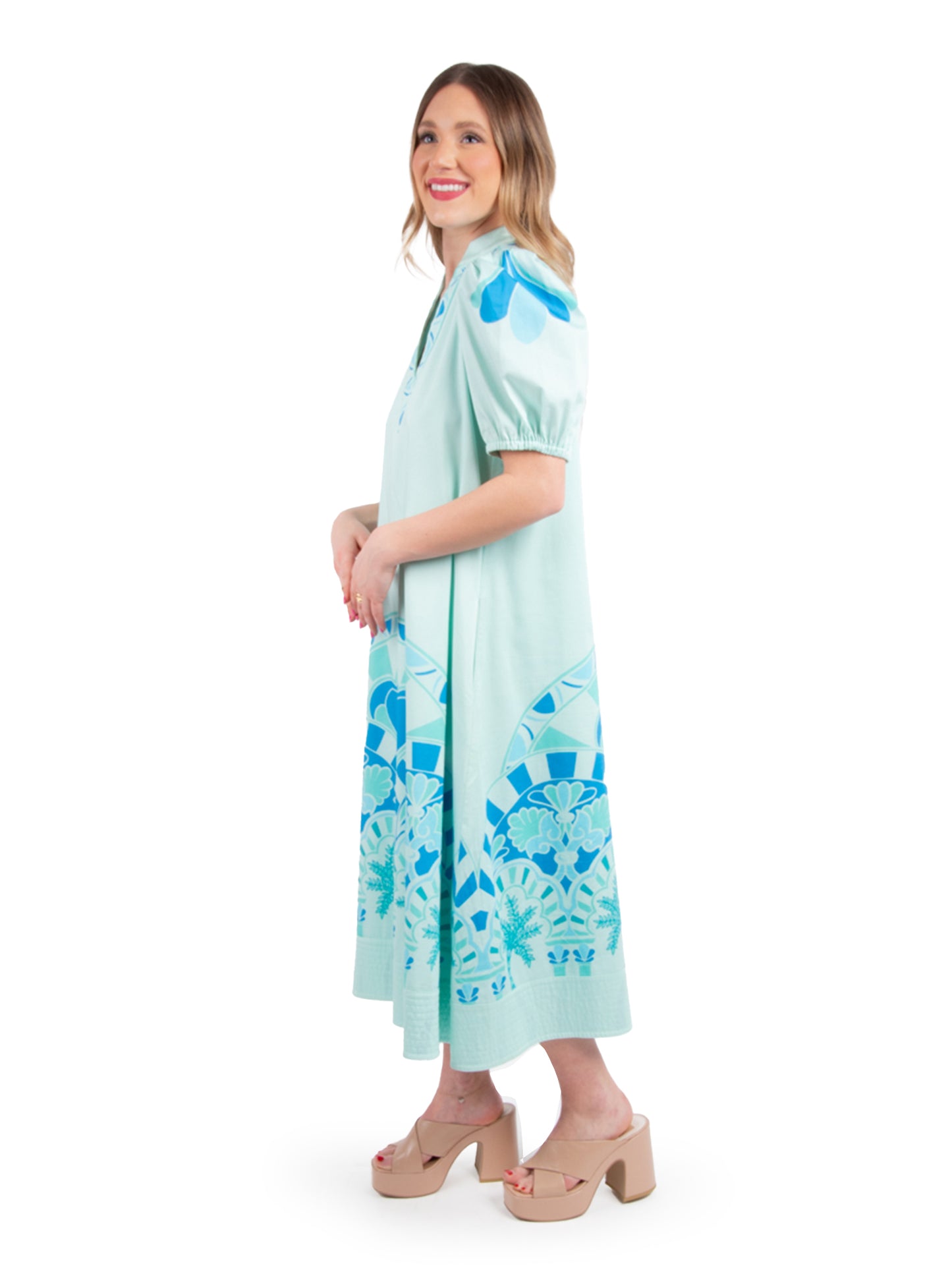 A woman beams in the Hampton Dress - Cordoba, featuring a light blue fabric adorned with bold floral patterns and charming puff sleeves. She stands elegantly in beige platform sandals against a plain white backdrop, showcasing versatile styling.