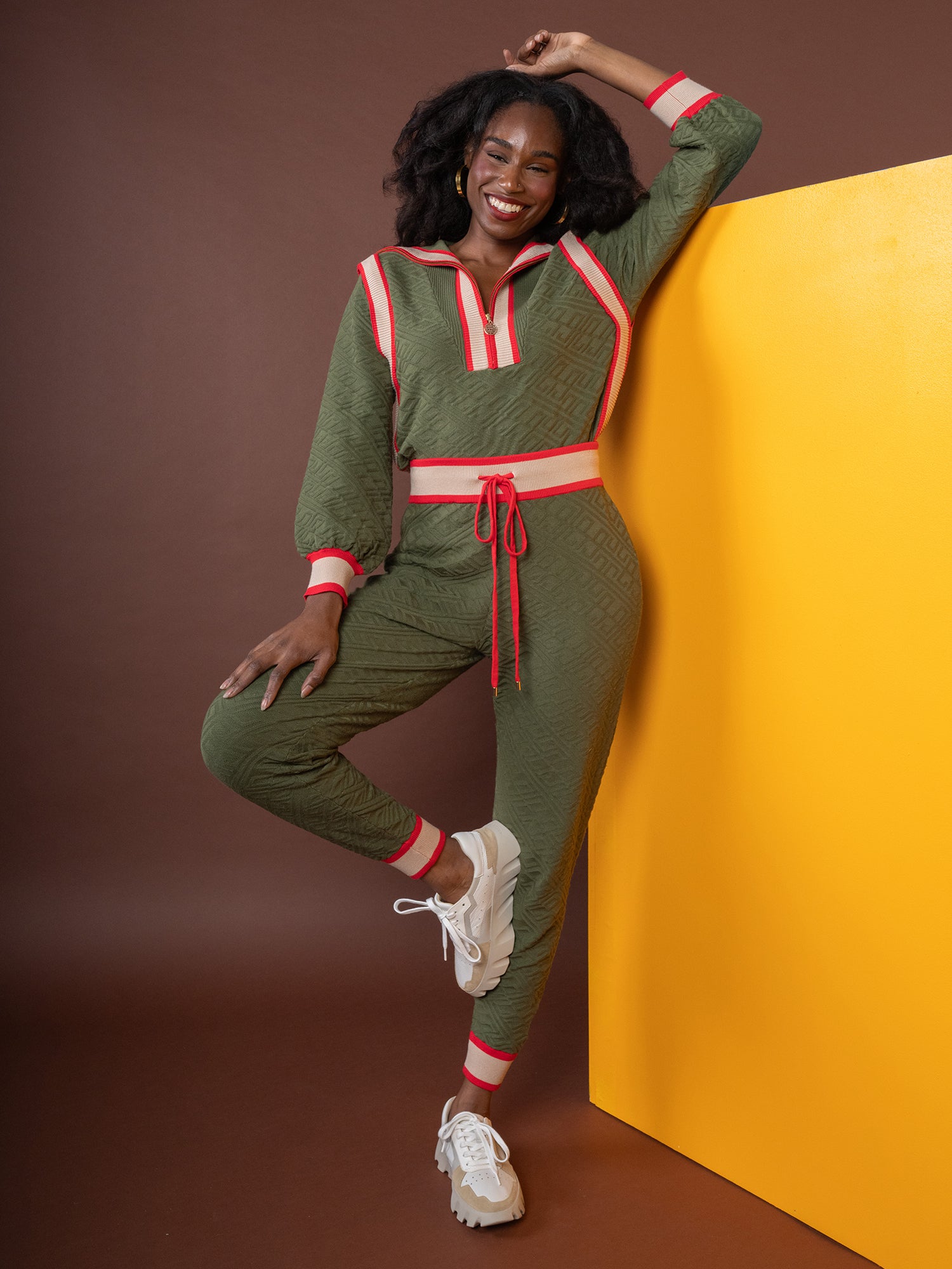 A woman in a chic Downtown Jogger - Olive Night Monogram jumpsuit with red and white trim stands smiling against a yellow panel. She wears white sneakers, capturing the essence of loungewear chic, with one hand on her head and the other on her hip. The background is brown.