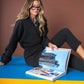 A person with long hair and glasses, dressed in a black Poppy Pop-Over - Jet Black Cheetah outfit, sits on a blue surface against a brown backdrop. They are looking at an open book featuring images of buildings and a beach scene.