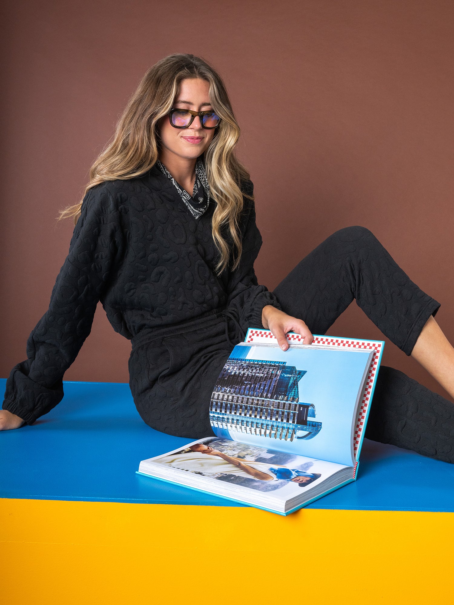 A person with long hair and glasses, dressed in a black Poppy Pop-Over - Jet Black Cheetah outfit, sits on a blue surface against a brown backdrop. They are looking at an open book featuring images of buildings and a beach scene.