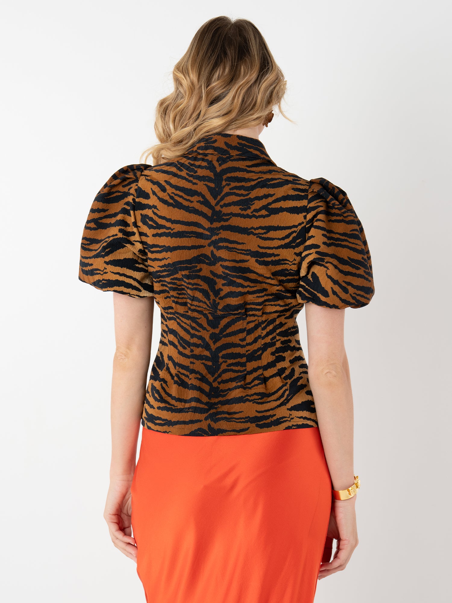 A woman with wavy blonde hair is seen from behind, wearing the Britt Top—a puff sleeve style with a black and brown print—paired with a bright orange skirt and accessorized with a gold bracelet on her right wrist.