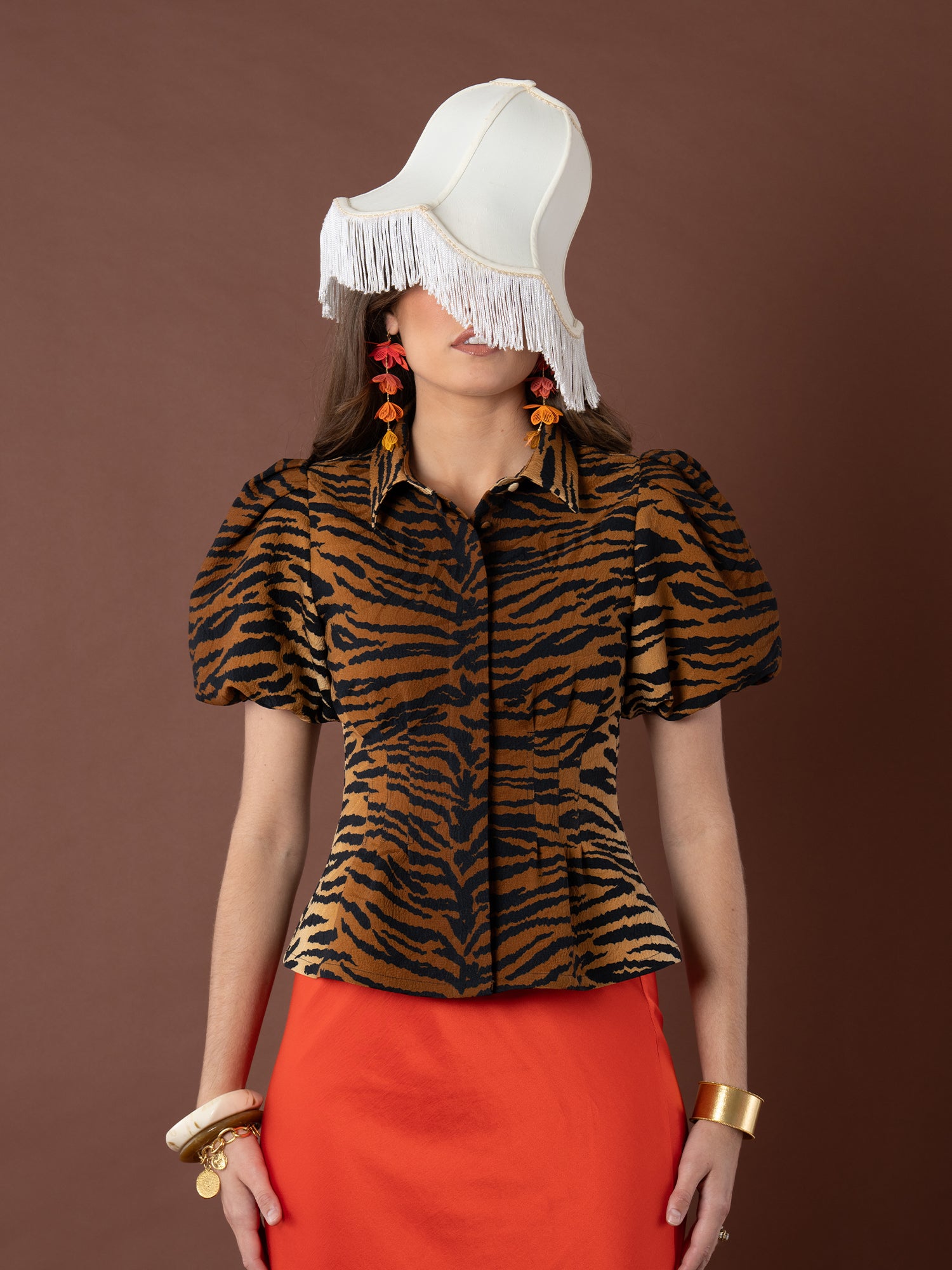 A woman in the Britt Top with puff sleeves, a bright orange skirt, bold jewelry, and a unique white fringe hat stands against a plain brown background.