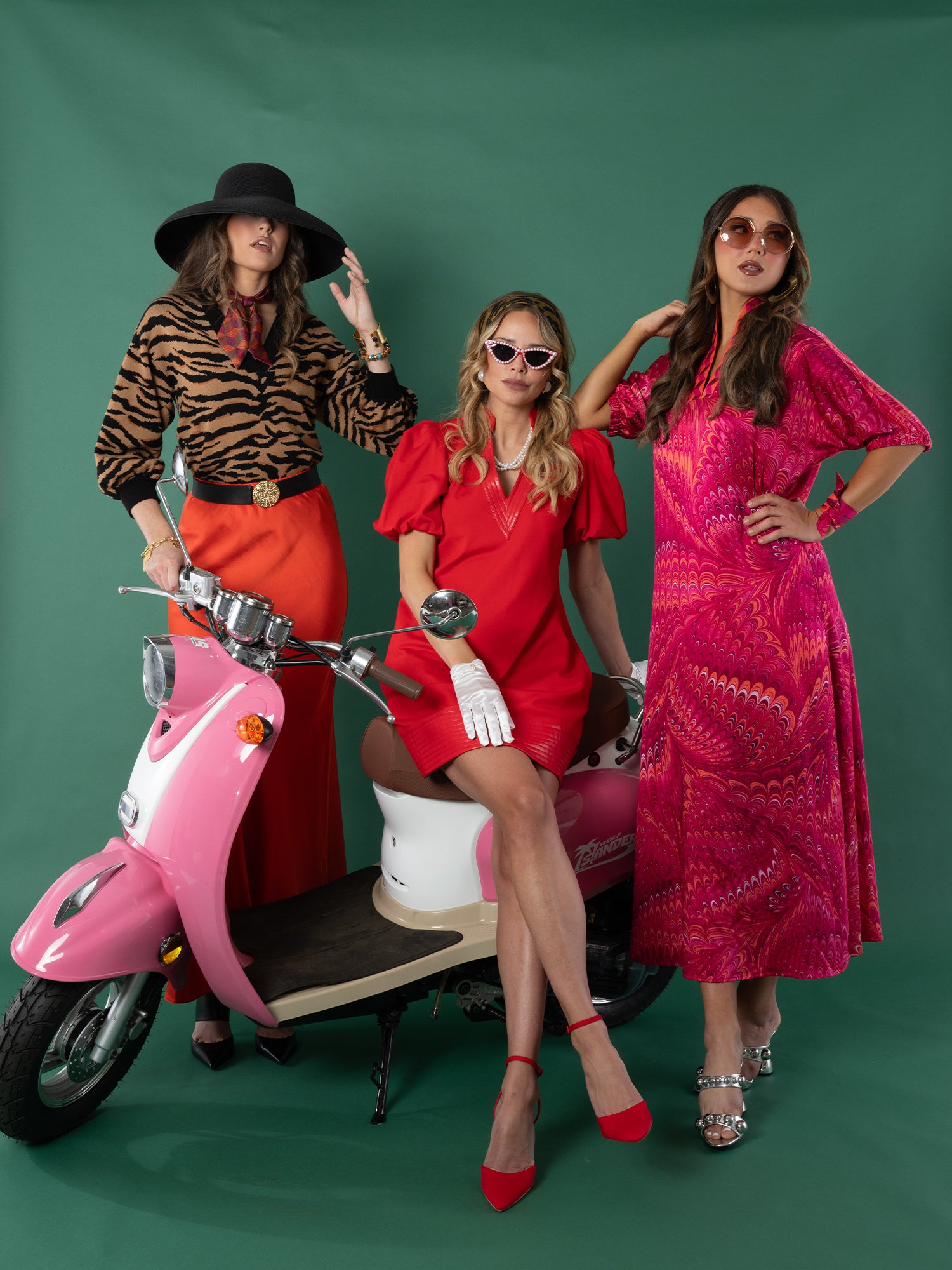 Three women exude vintage charm by a pink scooter: one in the Lolli Sweater and red pants, another in a red dress, and the third in a pink print dress with sunglasses—all set against a lush green backdrop.