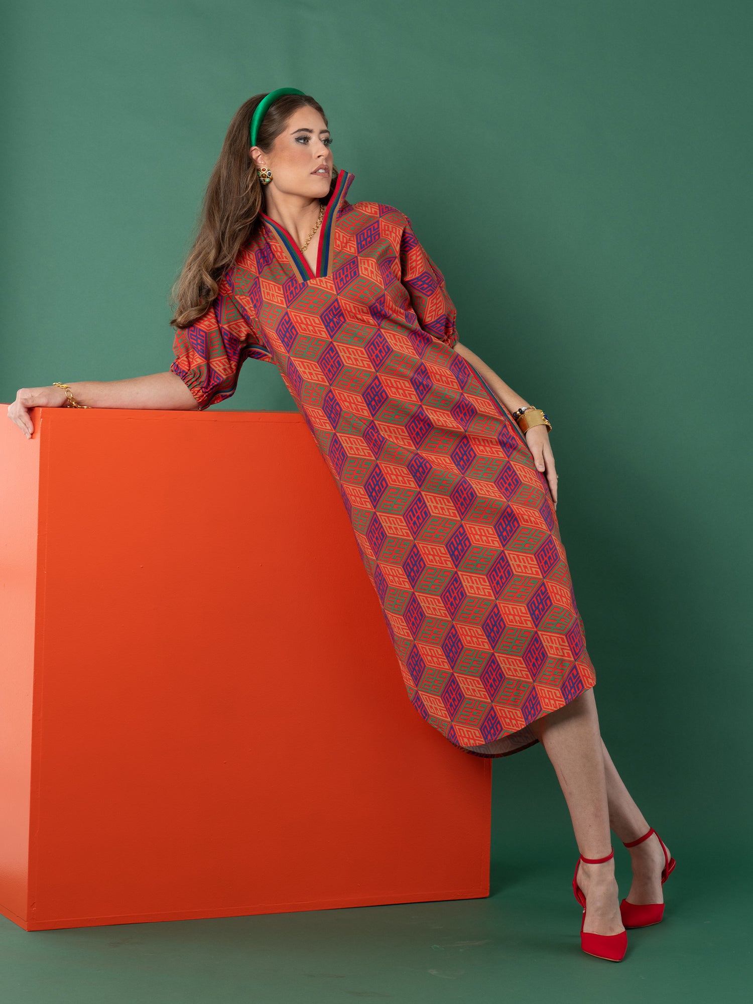 A woman in the Poppy Caftan, featuring a colorful geometric monogram print, leans against an orange block with a green backdrop. She pairs the dress with red heels, gold jewelry, and a green headband, her long hair worn loose.