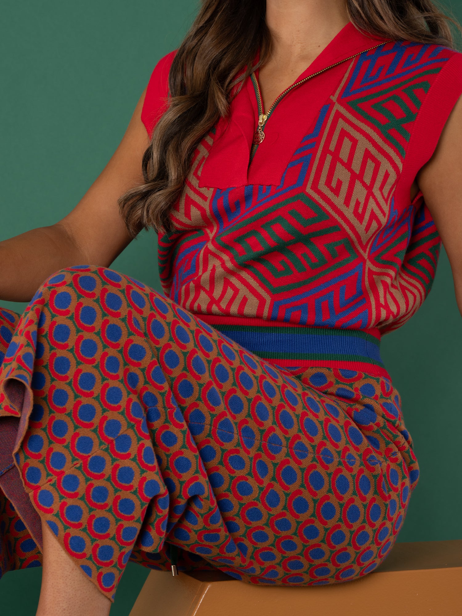A woman with long, wavy hair sits against a green background wearing the Poppy Pullover Vest—red, sleeveless, with geometric patterns—and high-waisted, wide-leg pants in a red, blue, and tan circular pattern.