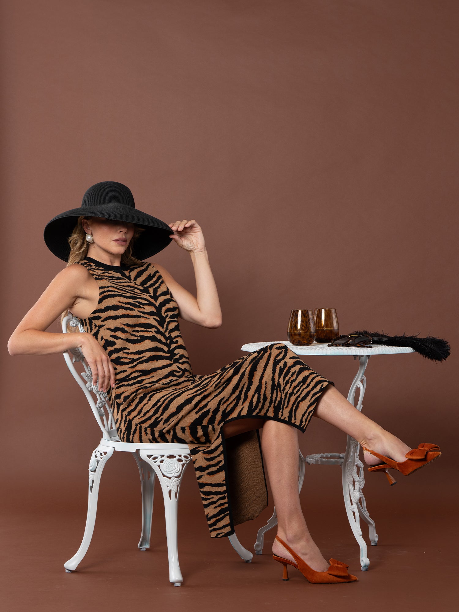 A woman wearing the Simone Sweater Dress and a large black hat sits at a small white table with two brown glasses, one arm resting on the table and her legs crossed, set against a brown background.
