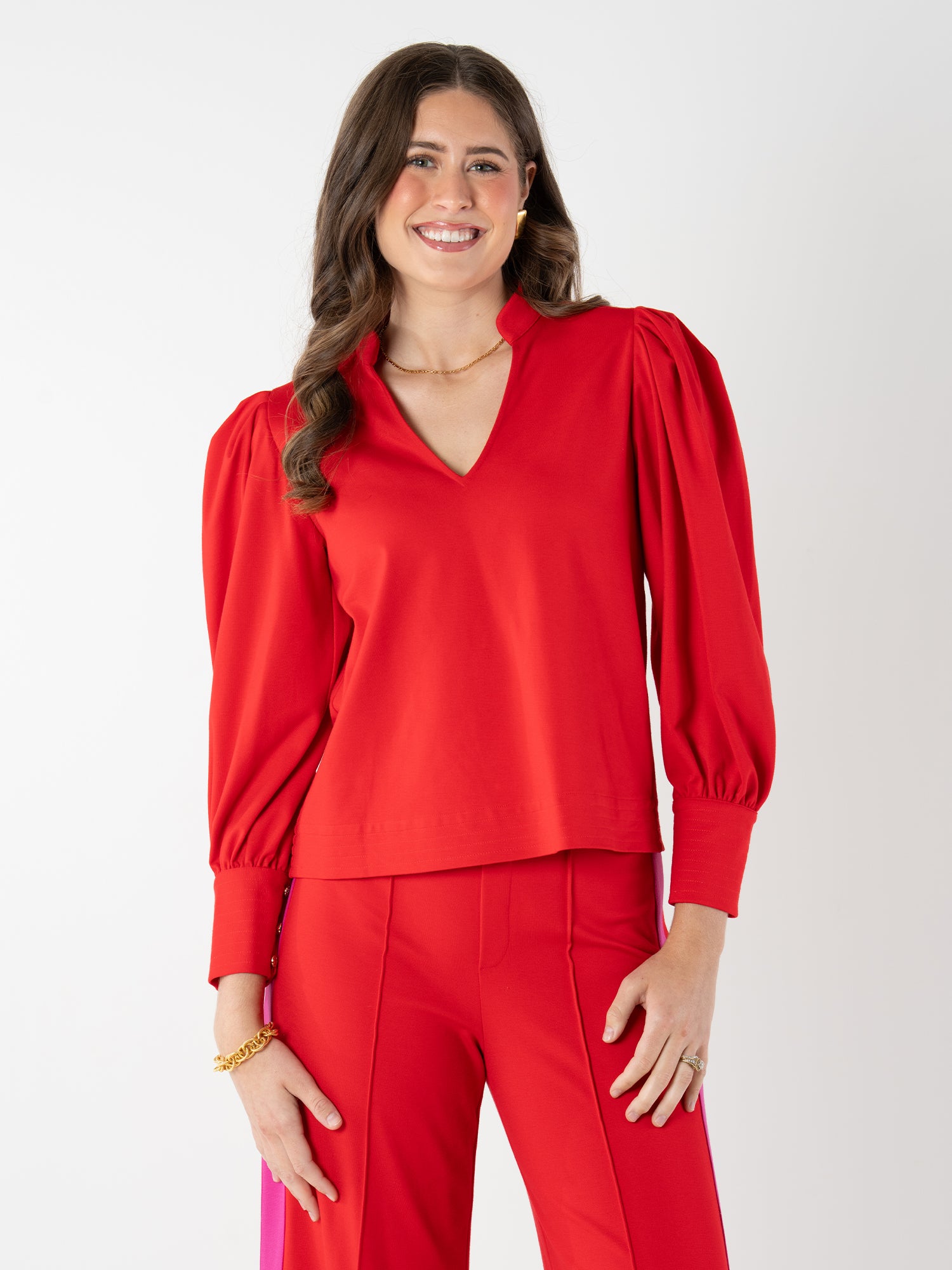 A woman with long brown hair smiles in a bright red Hampton Top with puffed sleeves and matching ponte knit pants, accessorized with gold jewelry, standing against a plain white background.