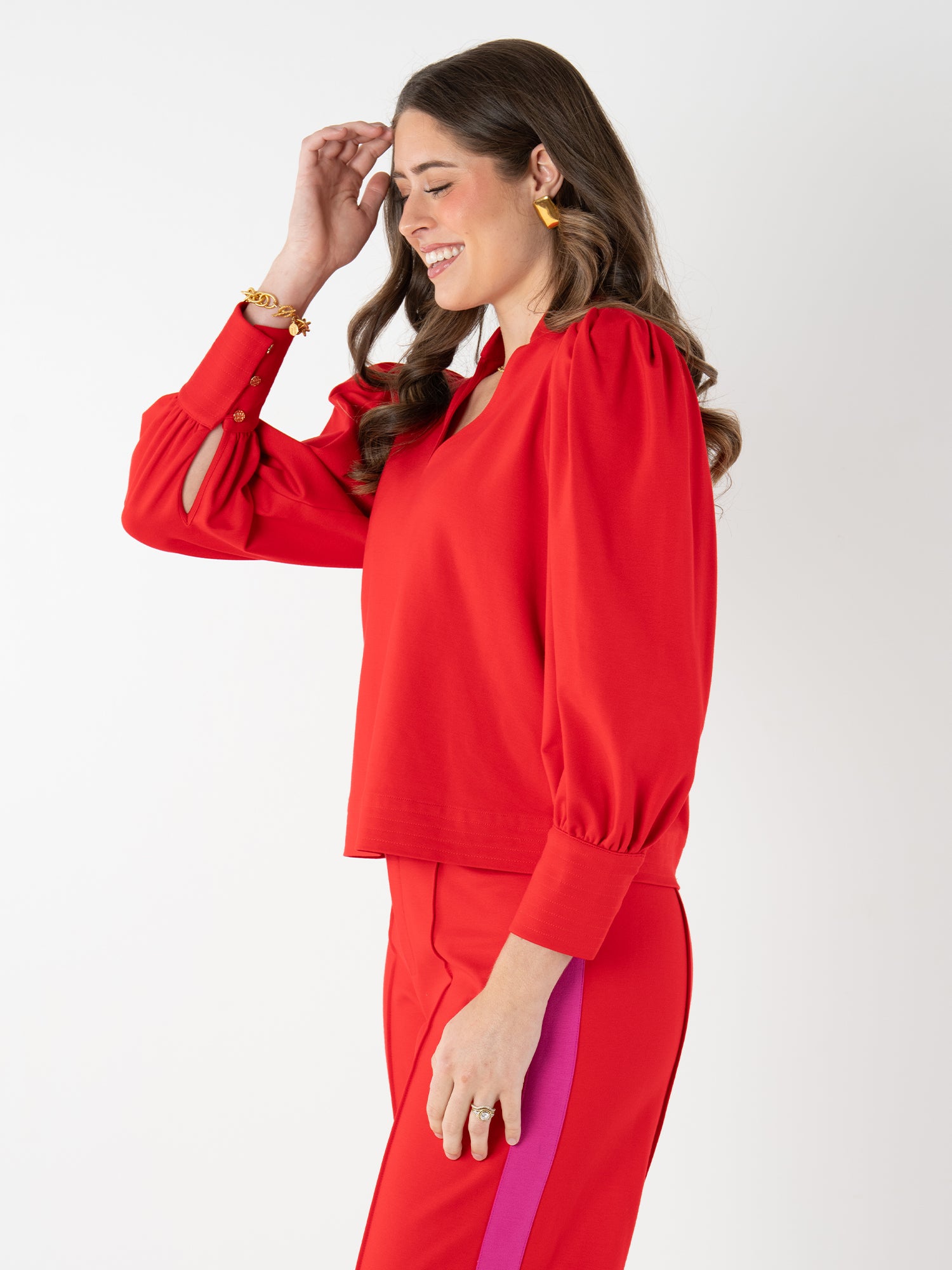 A woman with long brown hair smiles, touching her forehead. She wears the bright red Hampton Top with puff sleeves and matching red pants featuring a pink stripe, standing against a plain white background.