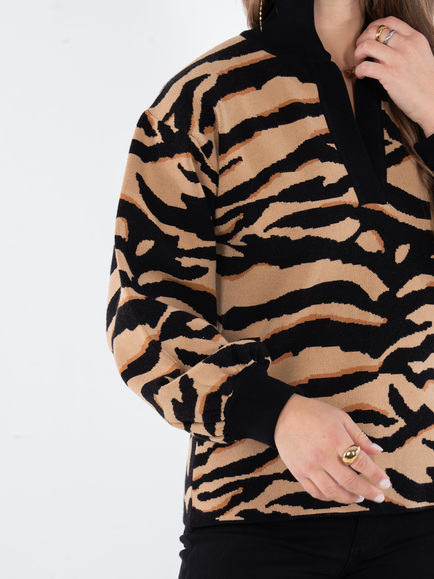 A person wears the Lolli Sweater featuring black and tan Lady Tiger print and vintage charm, paired with black pants. They touch their hair with one hand while gold rings on both hands catch the light near their waist.