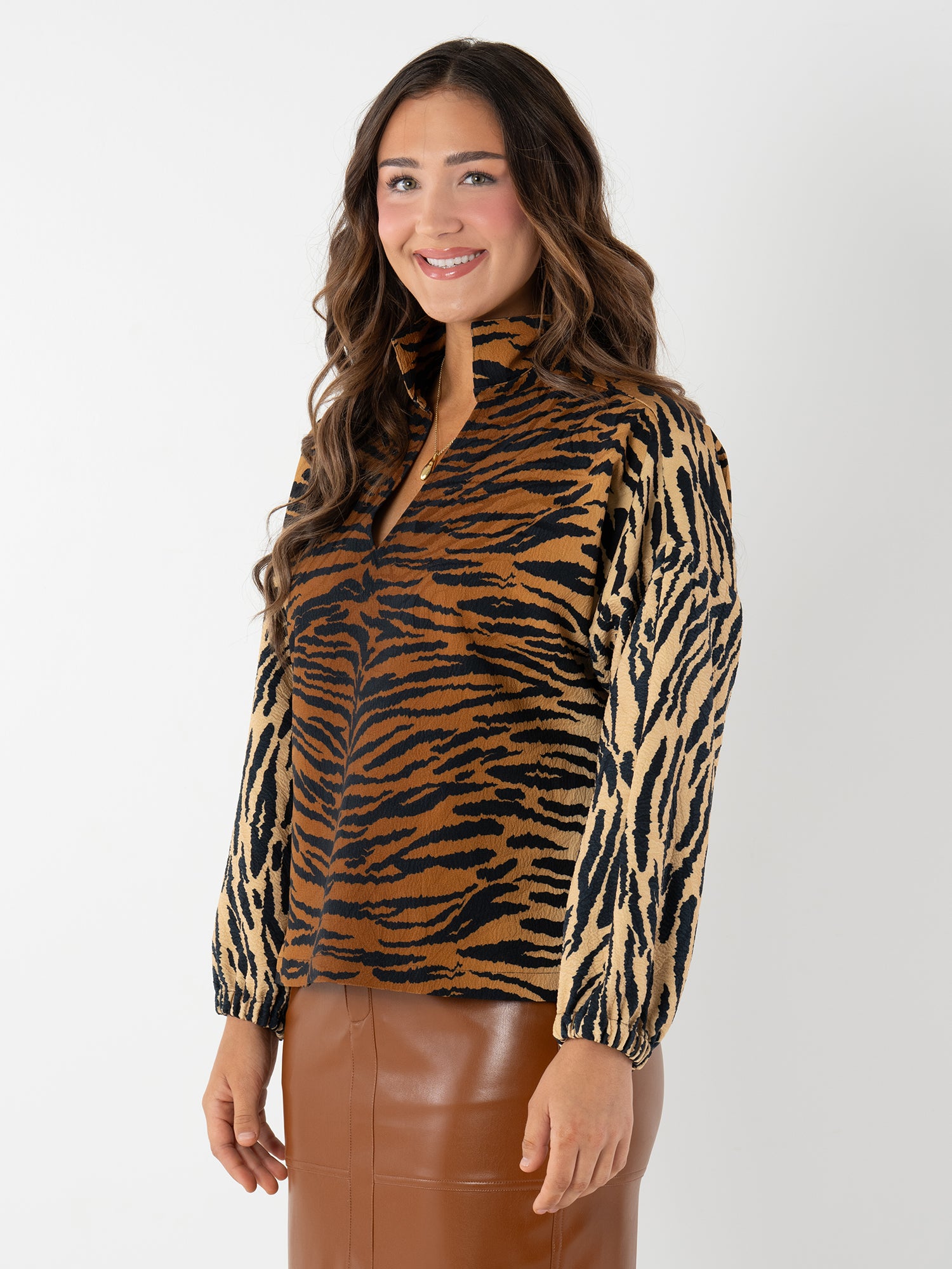A woman with long wavy brown hair smiles in front of a white background, wearing the Long-sleeve Poppy blouse with tiger jacquard print and a brown leather skirt.