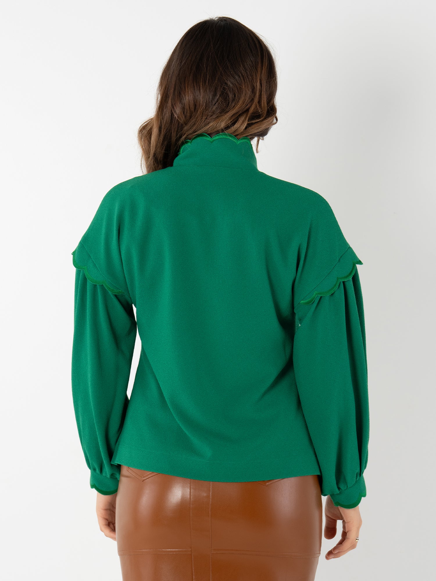 A woman with wavy brown hair is shown from behind, wearing the Orchid Top—a long-sleeved green blouse with blouson sleeves, ruffle accents, and a high collar—paired with a brown faux leather skirt.