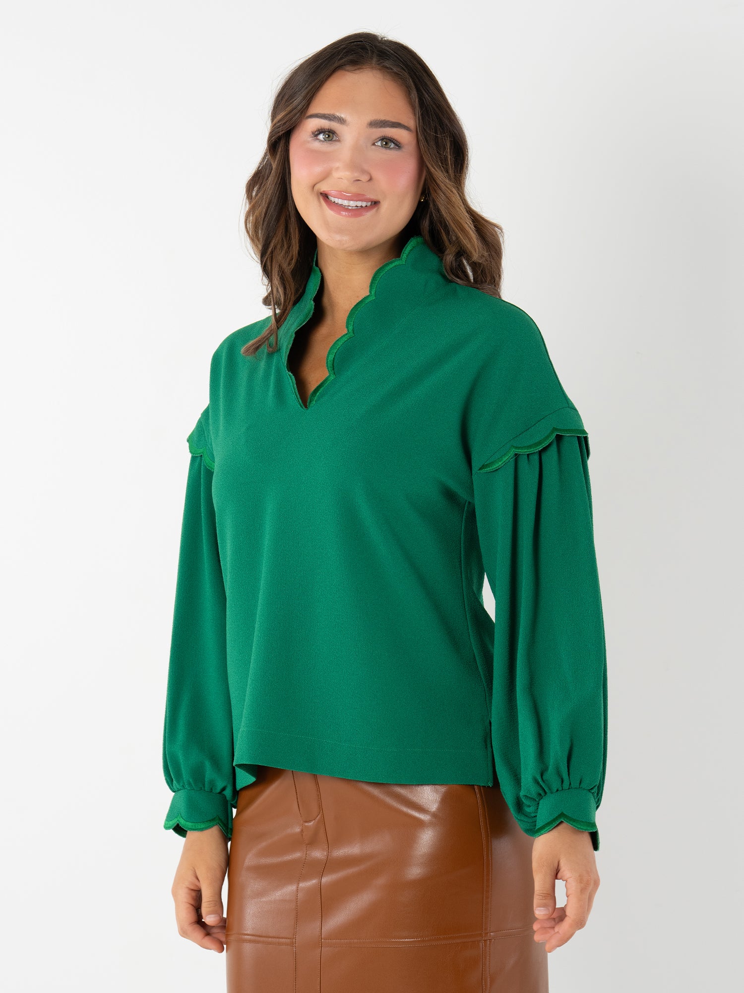 A woman with wavy brown hair wears the green Orchid Top, featuring blouson sleeves and a scalloped collar, paired with a brown leather skirt. She stands against a plain white background, smiling warmly.