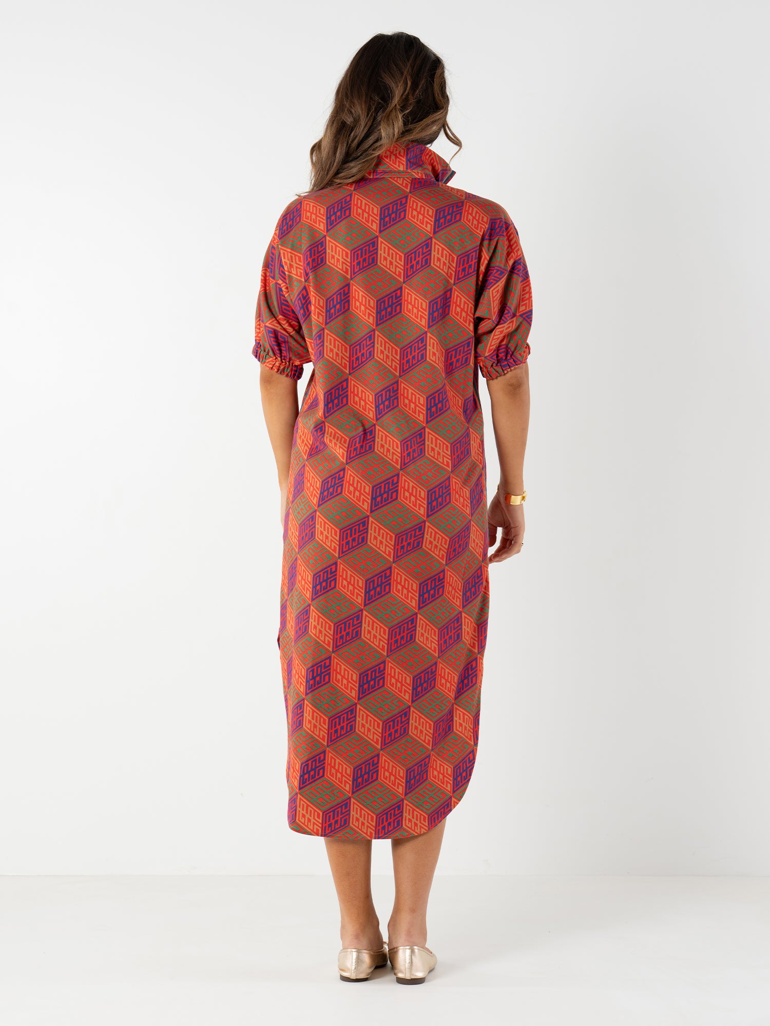 A woman with brown hair wears the Poppy Caftan, a mid-length dress featuring a red and purple geometric monogram print, elbow-length sleeves, and a high collar. She faces away and pairs it with light-colored flat shoes.
