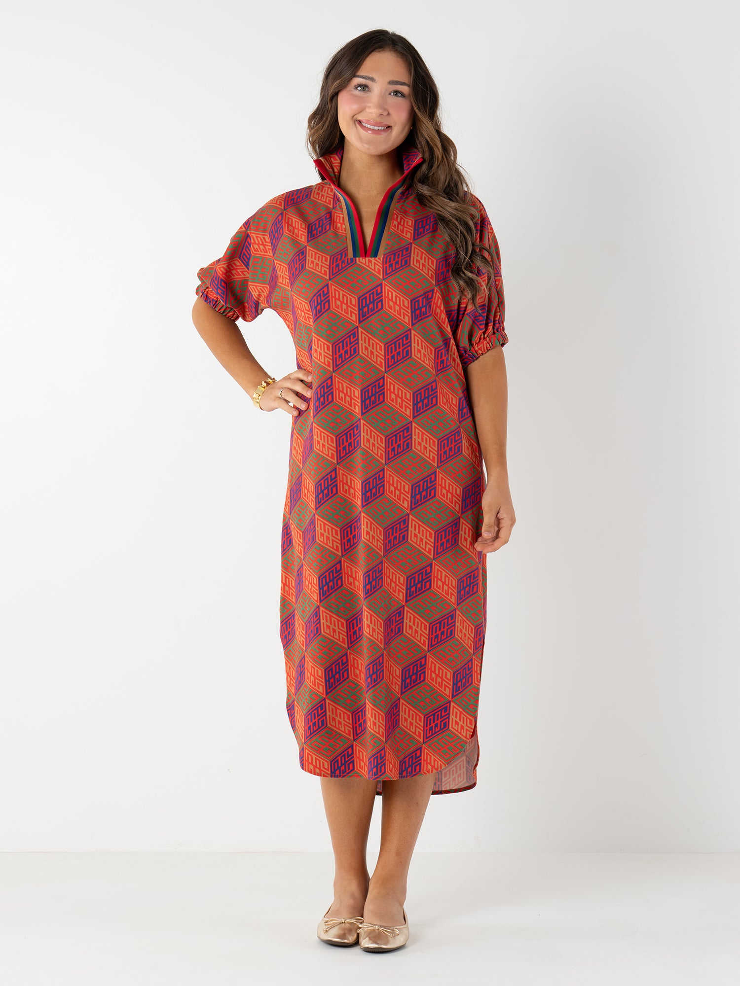 A woman with long, wavy hair smiles in a Poppy Caftan with a vibrant geometric monogram print, elbow-length sleeves, and a zip collar. She pairs it with beige ballet flats against a plain white background.